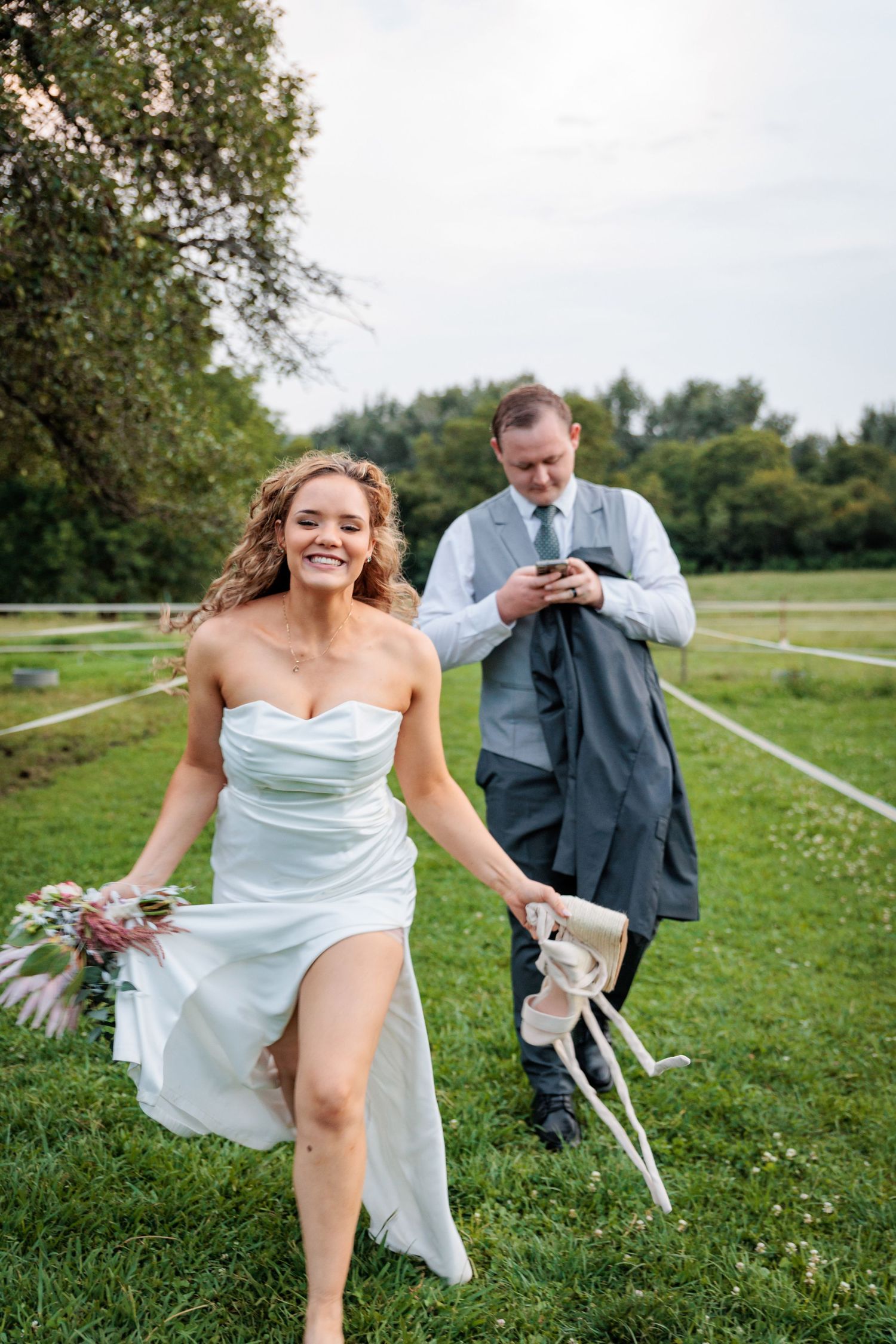 A playful wedding moment in a sunny meadow with rustic fencing.