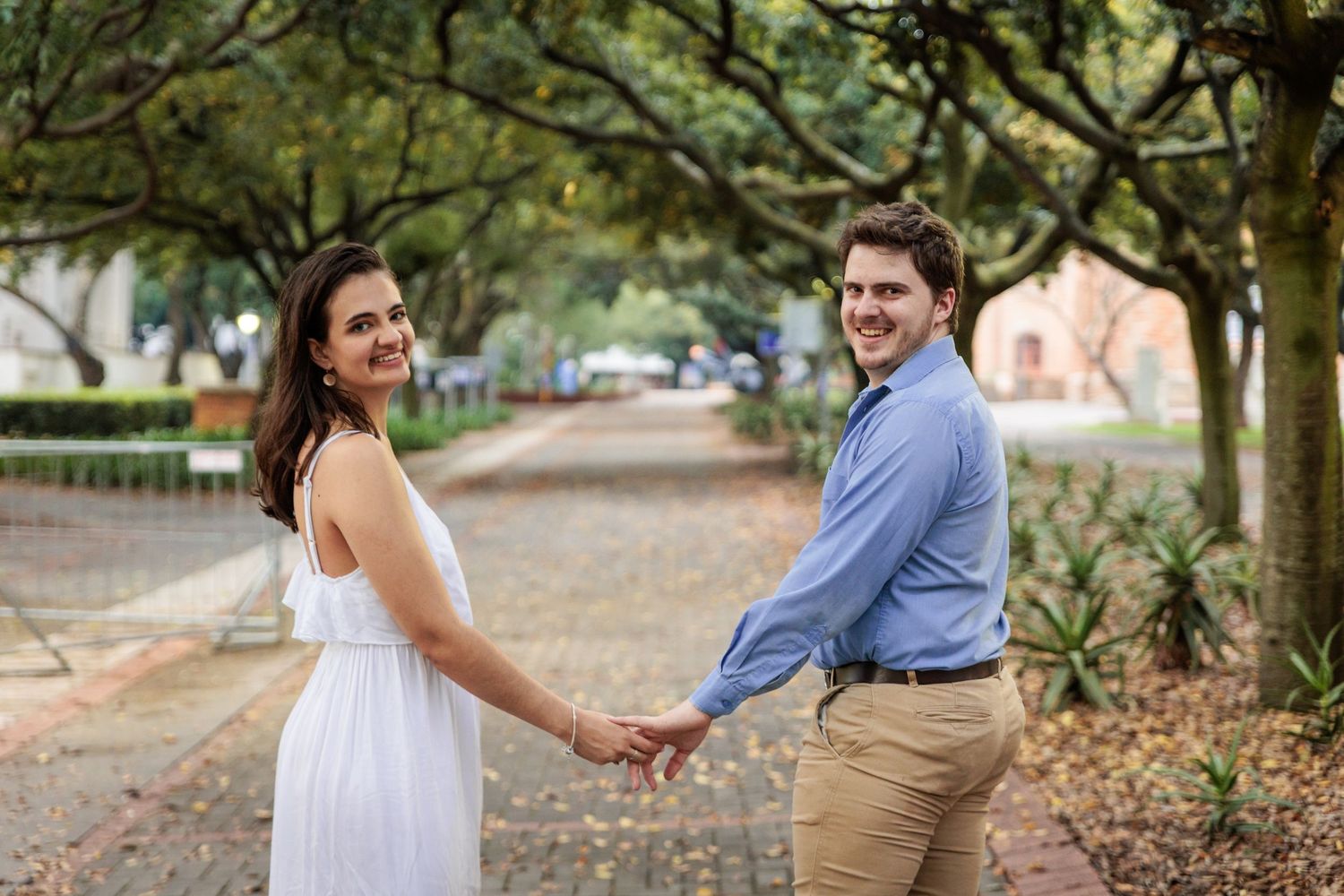 A romantic couple holds hands and walks through a lush garden setting during an engagement photo session.