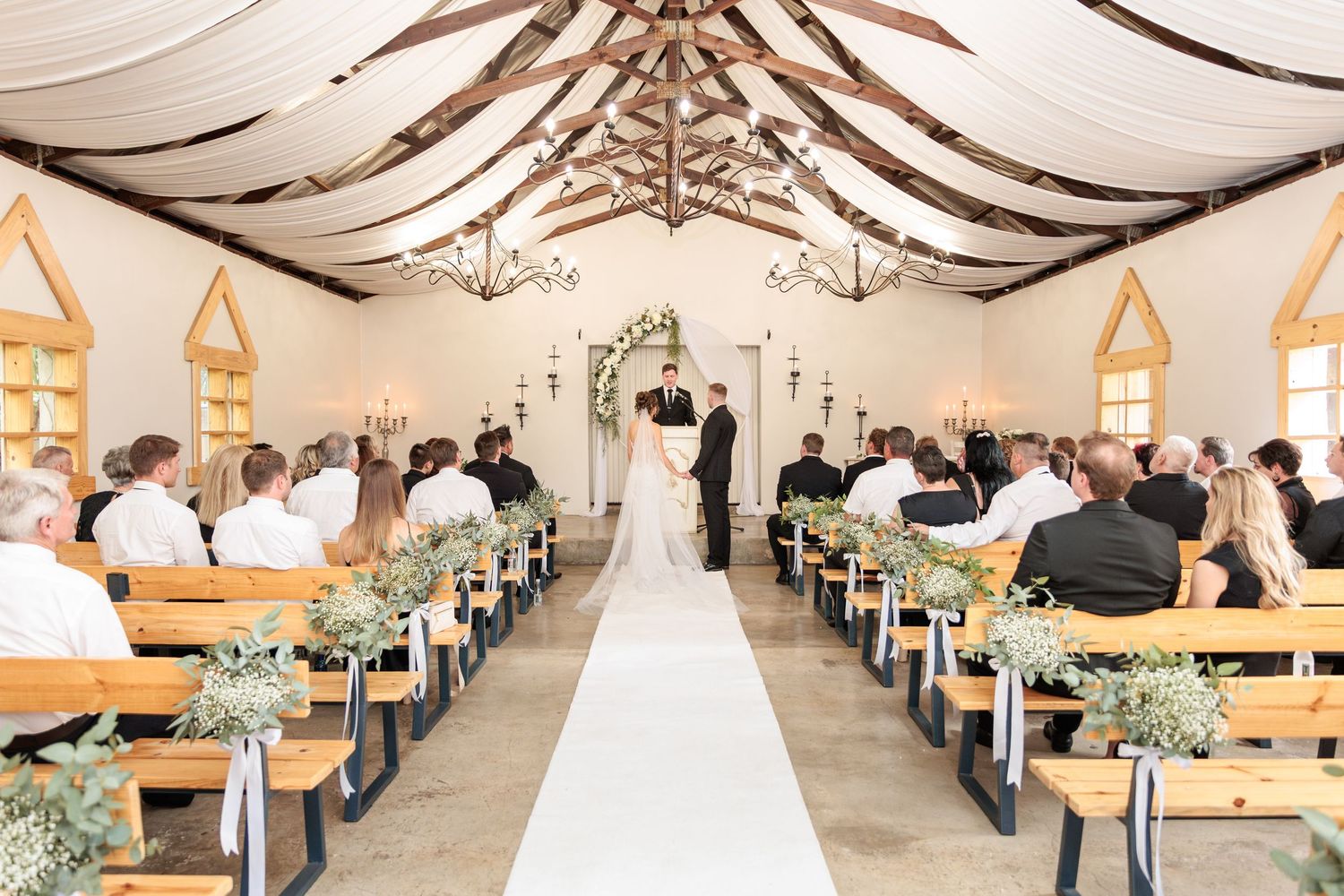 Wedding ceremony inside a rustic chapel with wooden pews, draped fabric ceiling, and floral decorations along the aisle.
