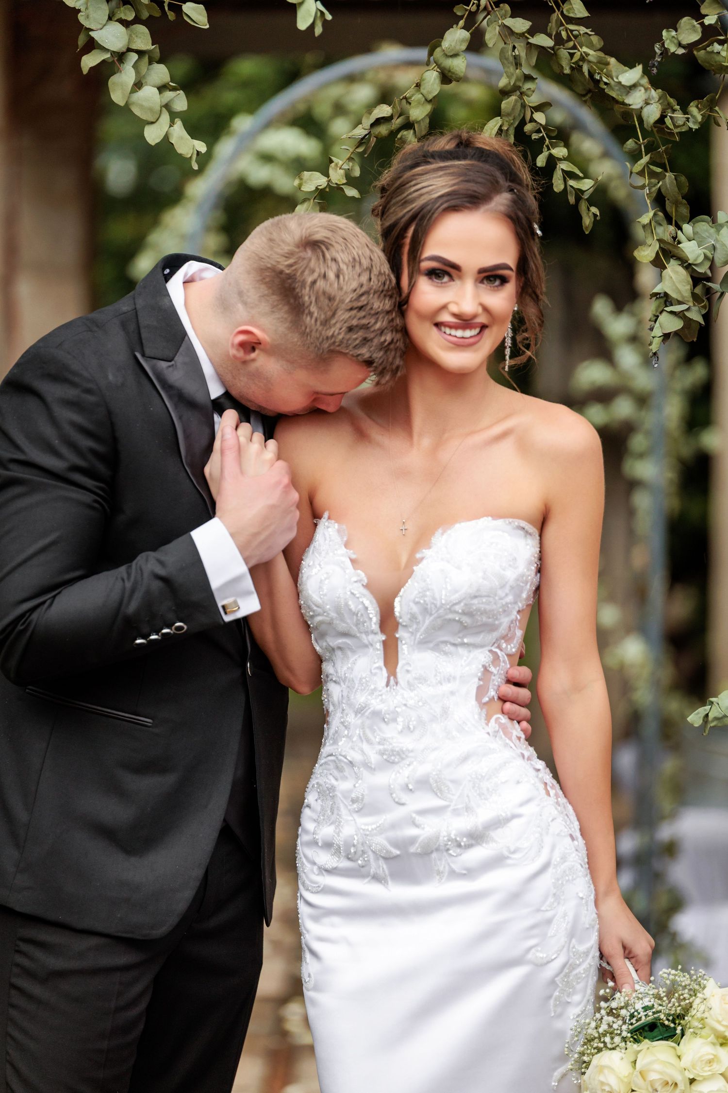 A romantic wedding moment captured under a floral arch with a bride in a fitted white lace gown and groom in black suit.