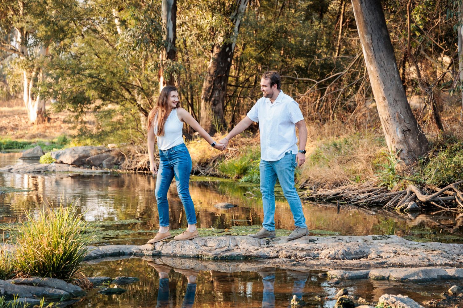 A couple in matching blue jeans and white shirts poses together by a scenic creek in a natural outdoor setting.