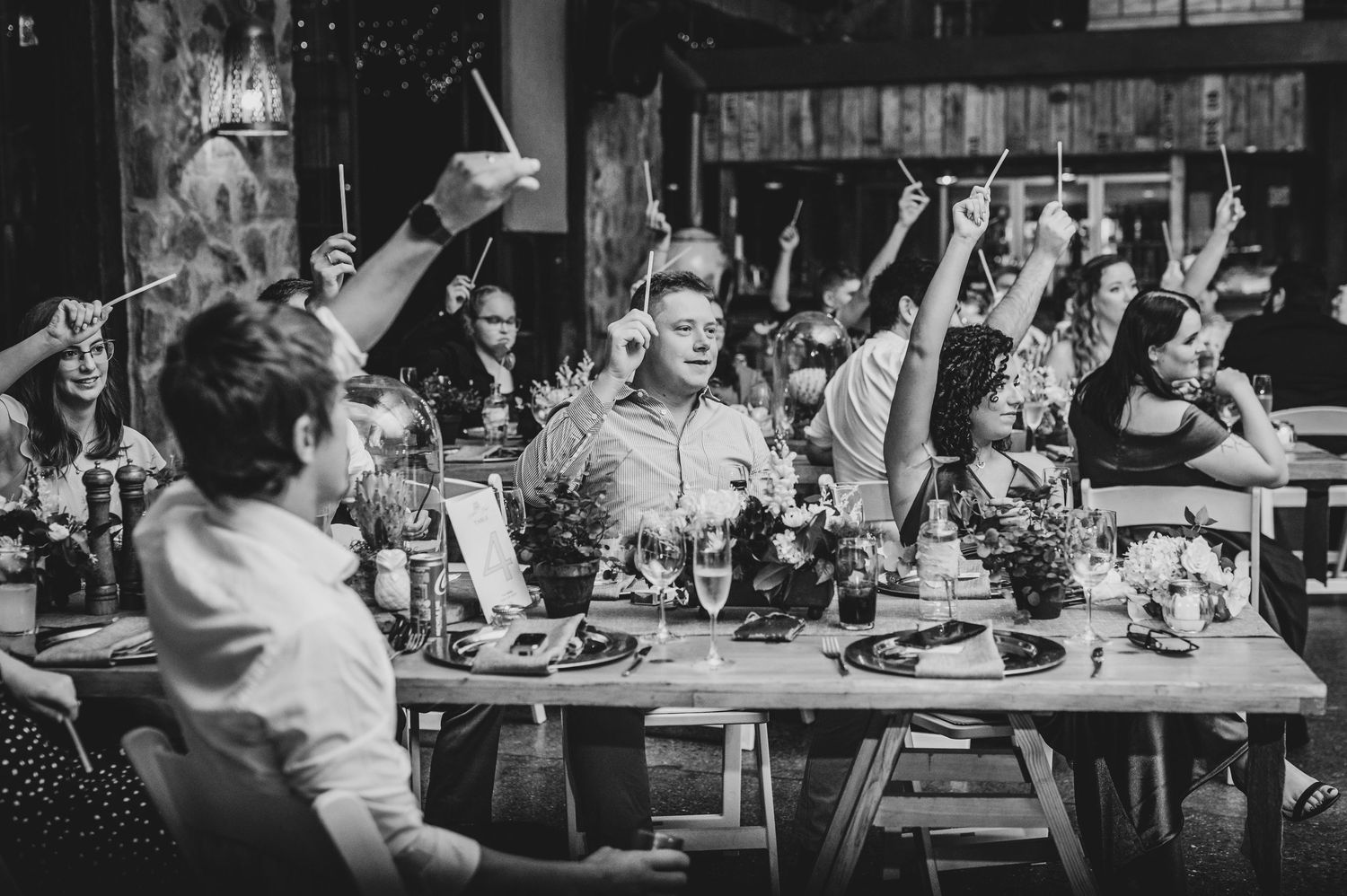 A black and white photo of wedding guests celebrating with raised arms at a reception dinner.