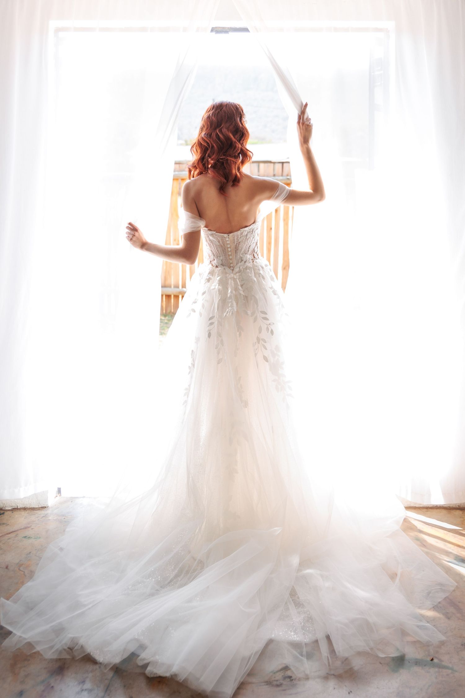 Ethereal bridal portrait with flowing white wedding dress against backlit window.
