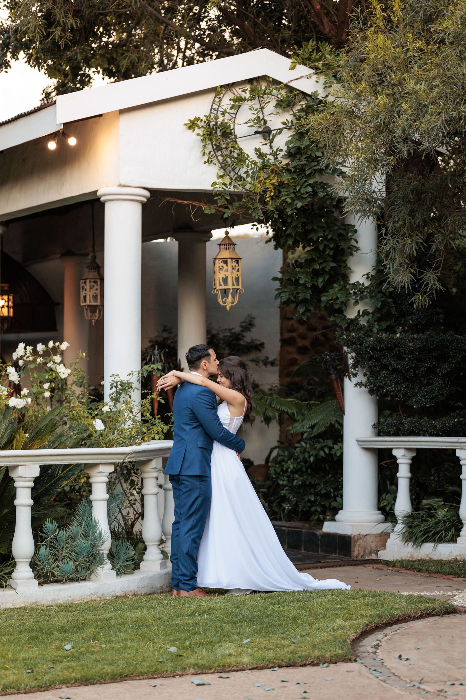 Wedding couple shares intimate moments along a white columned porch with greenery.