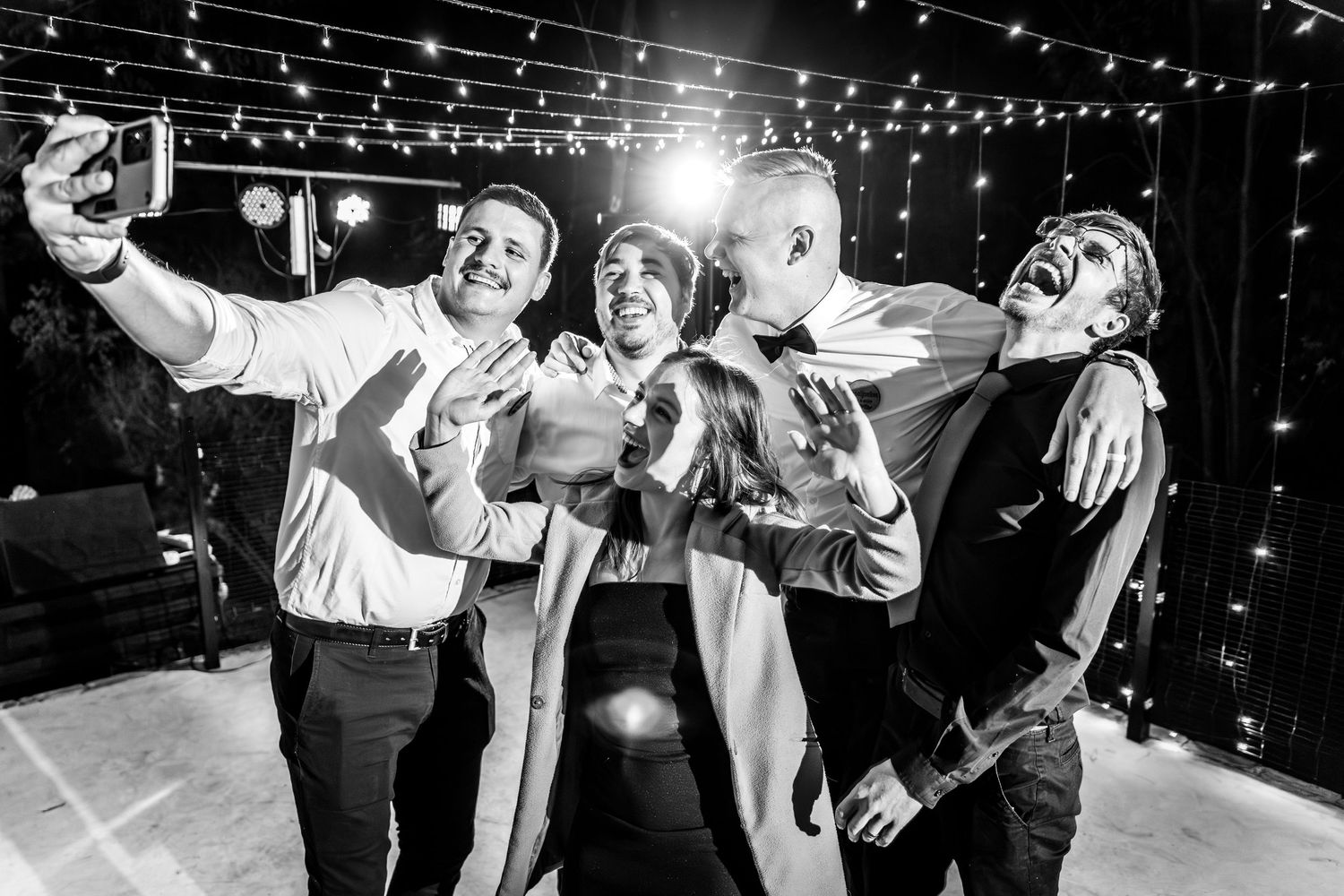 Black and white photo of four friends taking a fun selfie at a party with string lights in background.