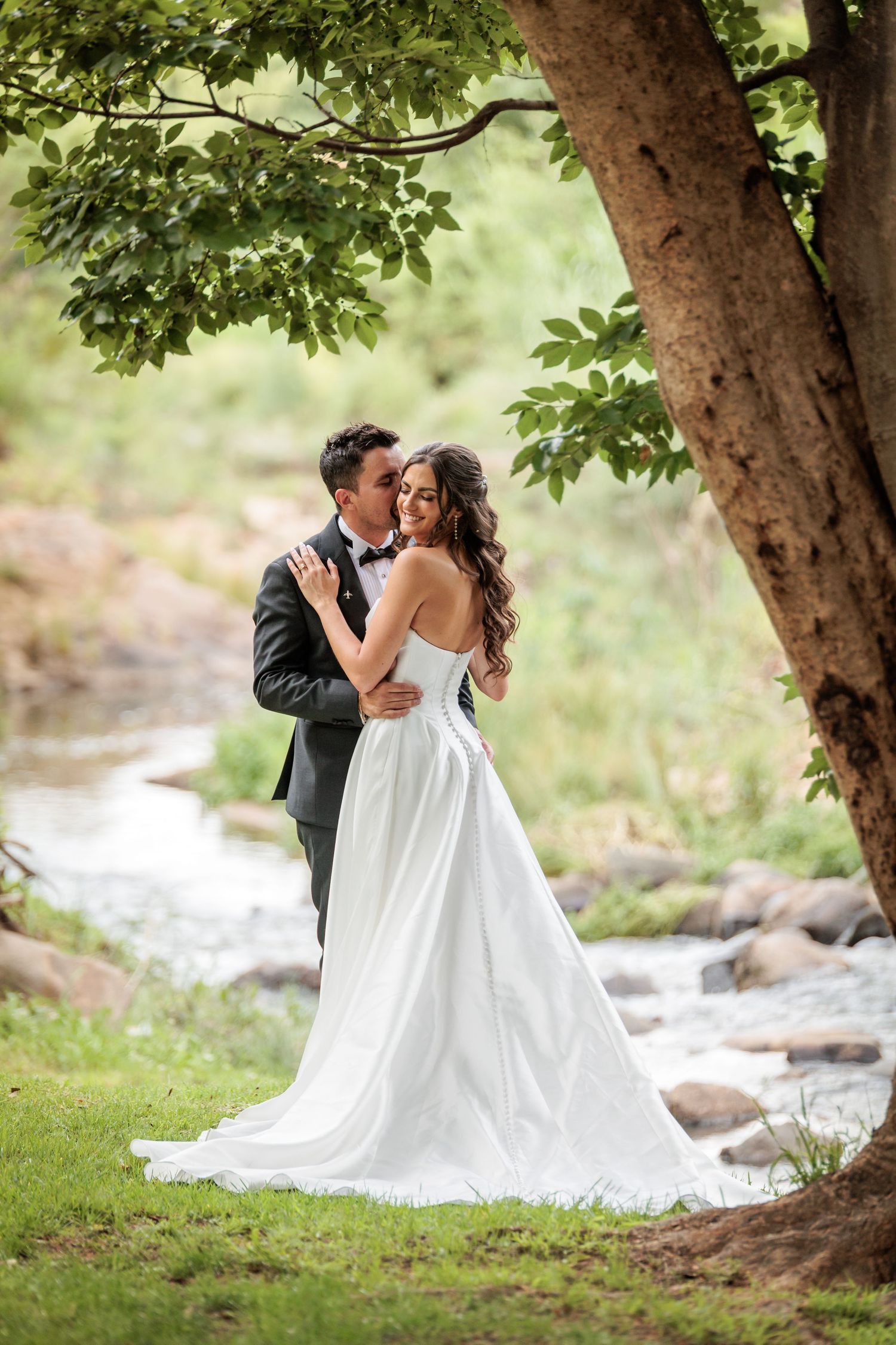 A romantic wedding couple embraces under a large tree beside a flowing stream in a dreamy outdoor setting.