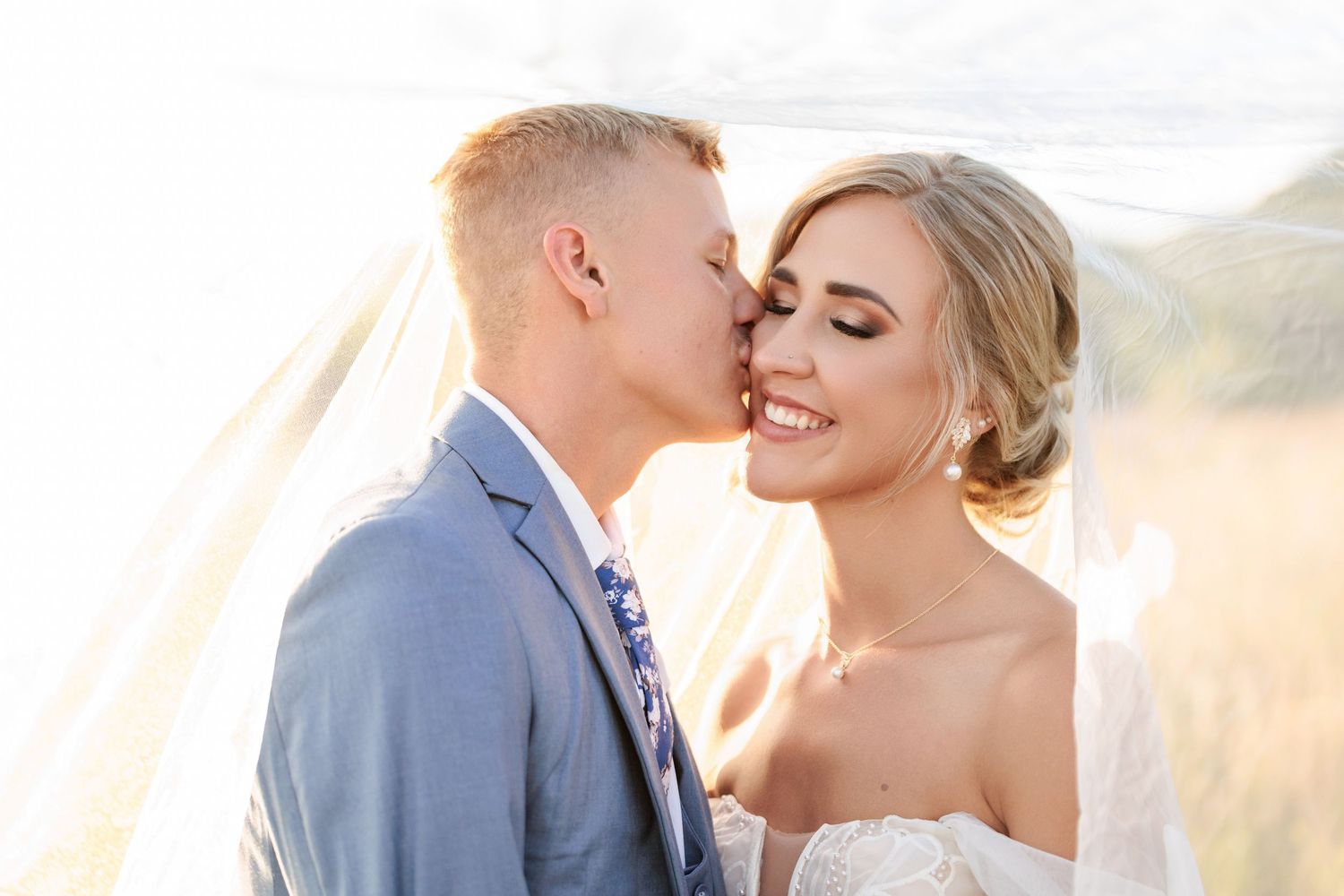 A romantic wedding couple embraces in a sunlit field during golden hour with mountains in the background.