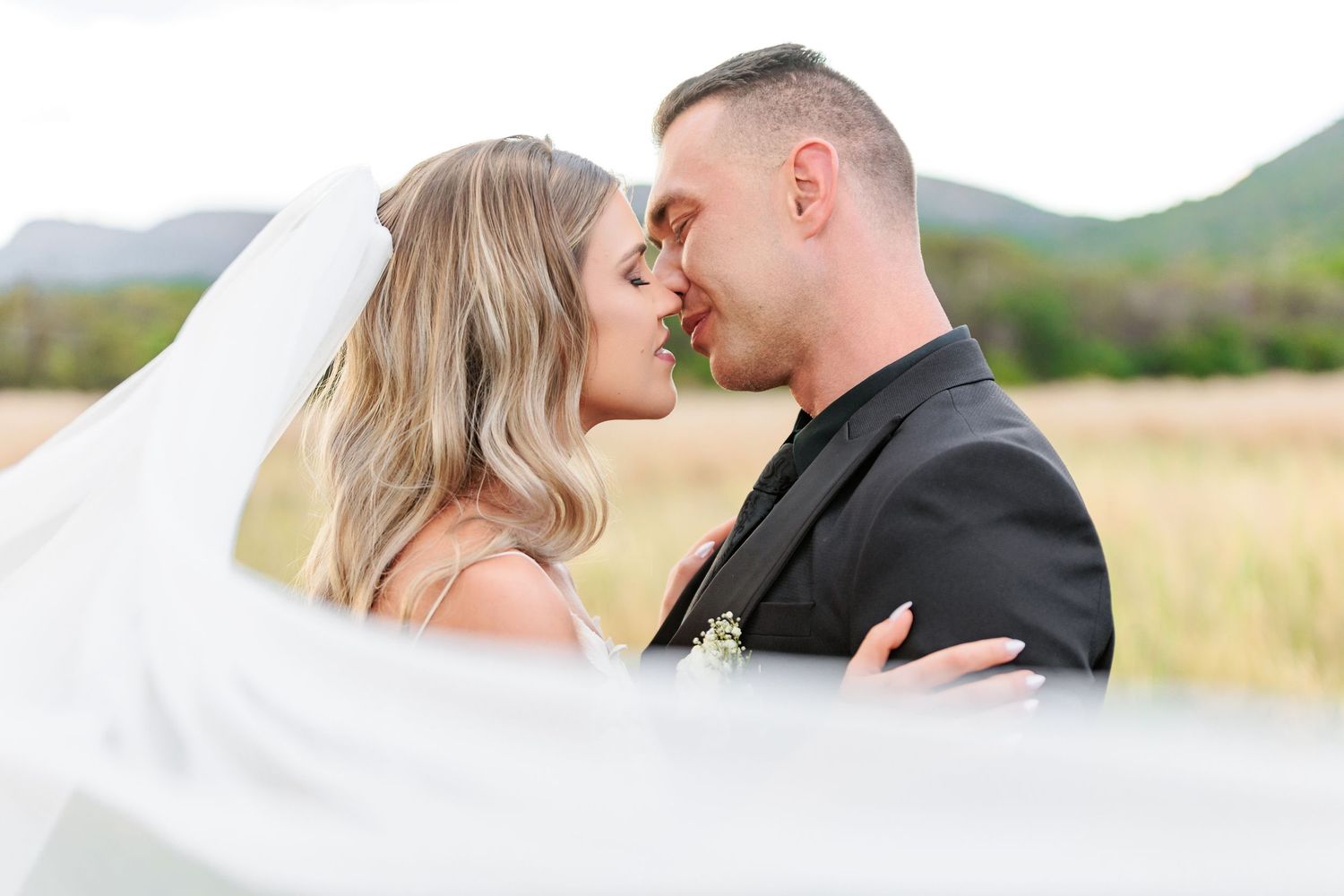A romantic wedding couple shares an intimate moment in a field with mountains in the background.