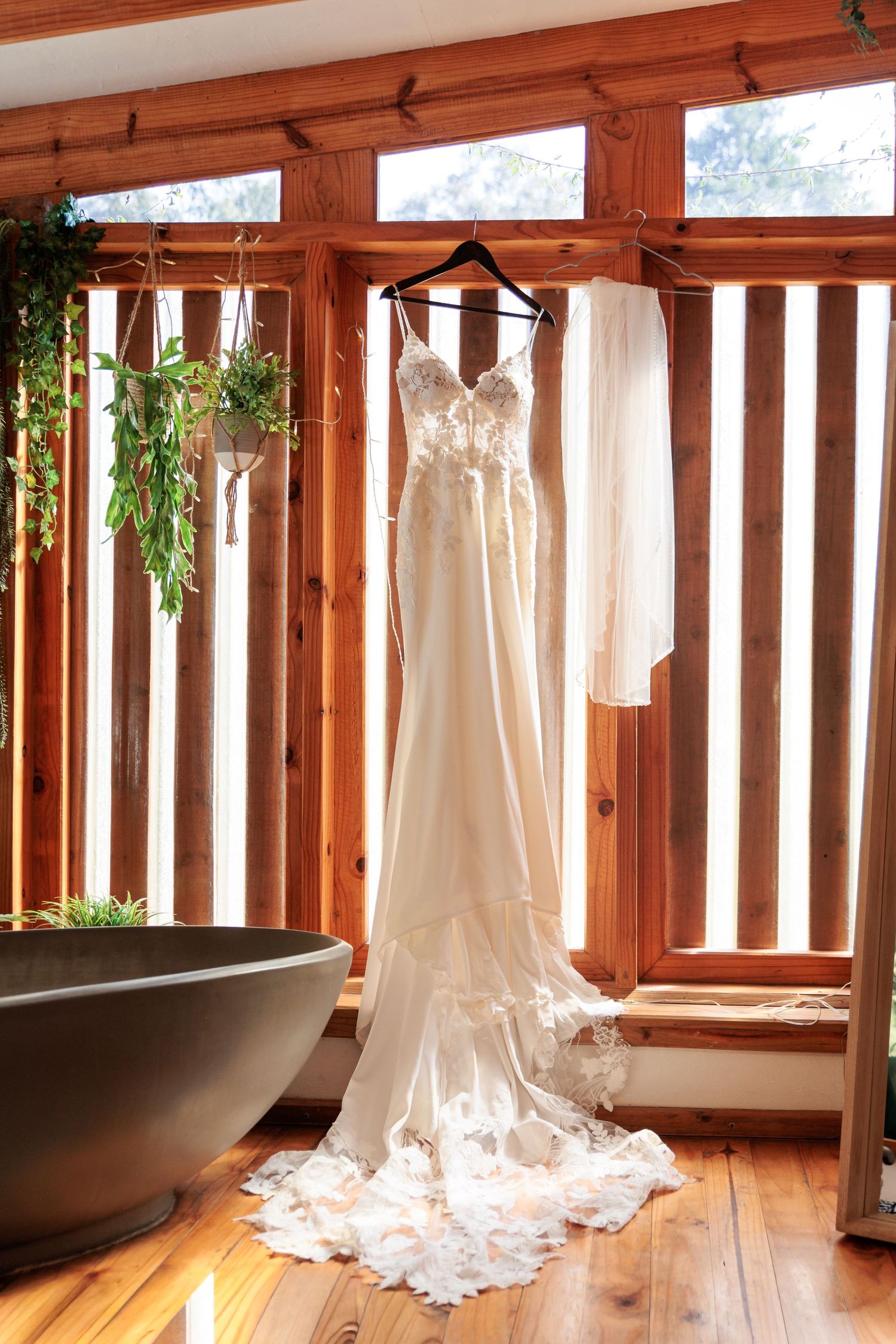 A wedding dress hangs elegantly in a sunlit wooden room with large windows and a freestanding bathtub.