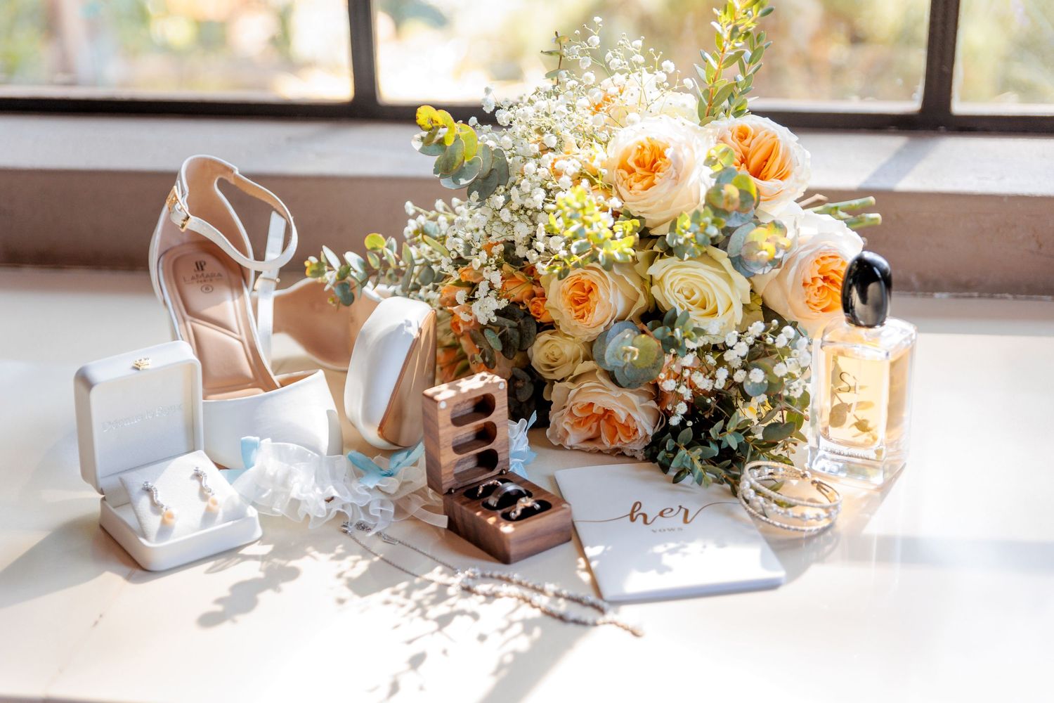 Elegant bridal flat lay with peach roses bouquet, gold heels, jewelry boxes, perfume, and a personalized notebook on white surface.