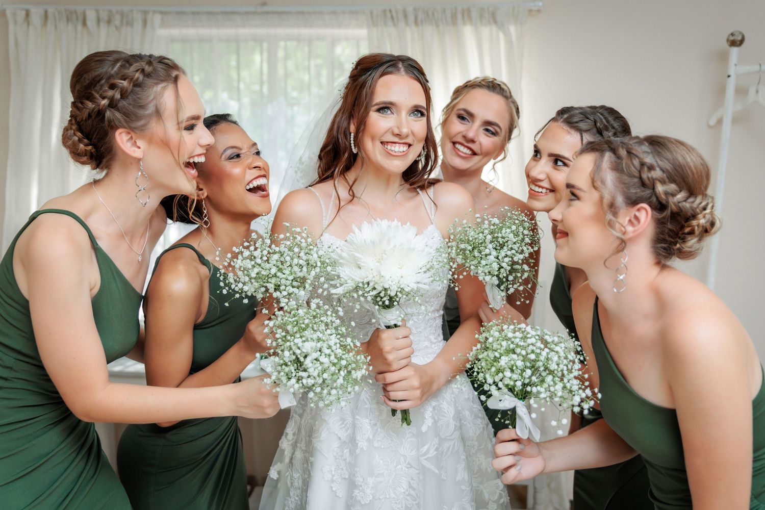 A wedding party in green bridesmaid dresses and white gown share joyful laughter together.