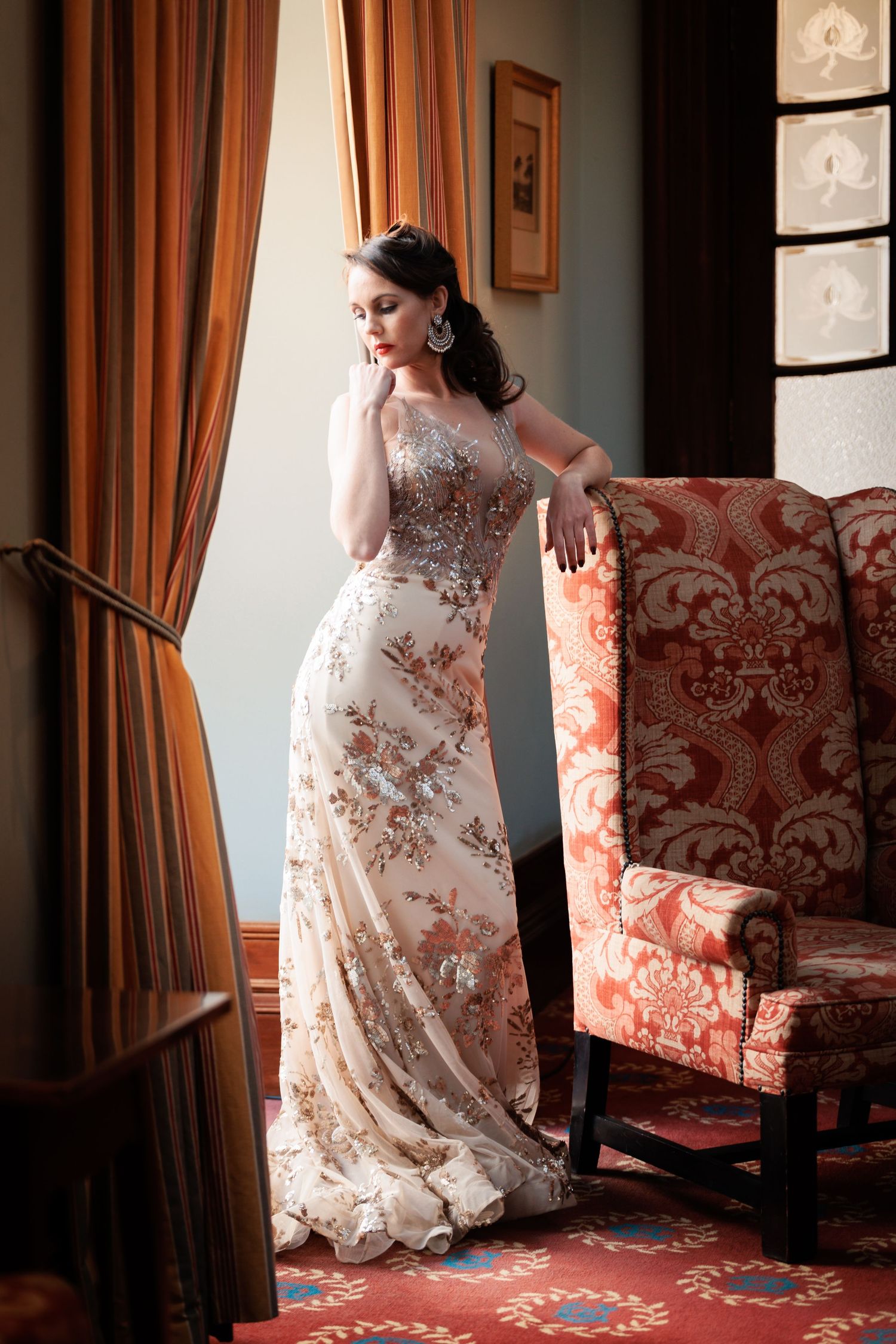 Elegant white lace wedding dress with floral details photographed against red damask chair and curtains in natural light.