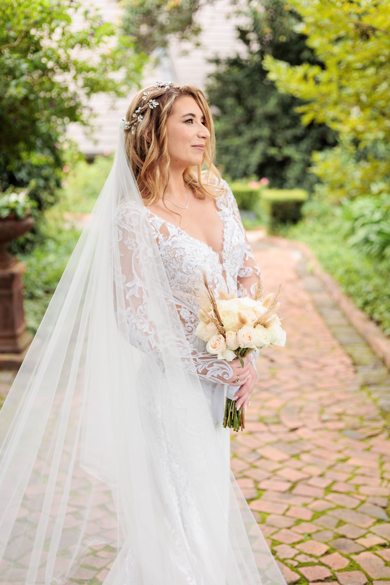 A bride in a white lace wedding gown with flowing veil poses in a garden setting with stone pathway and greenery.