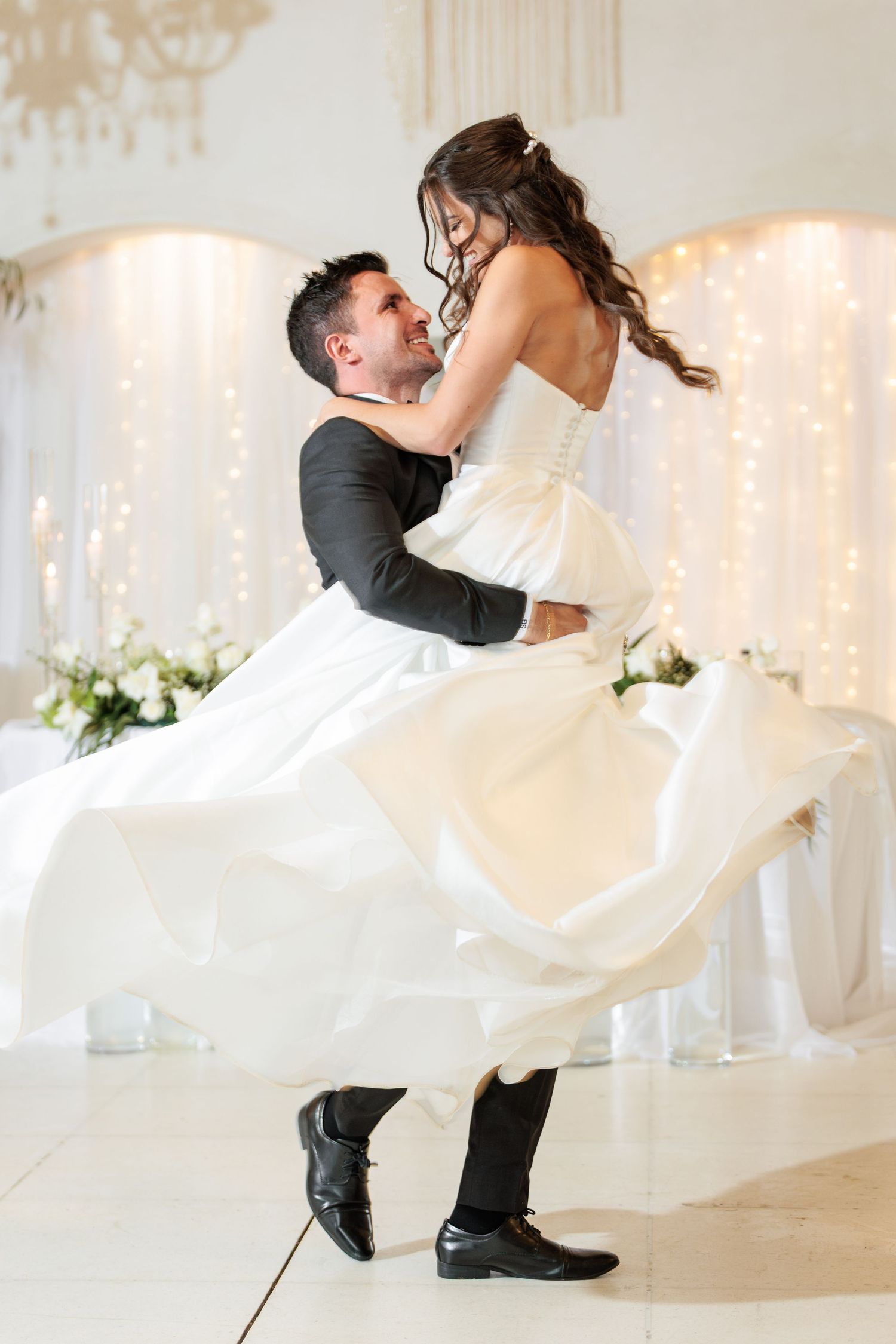 A couple in wedding attire performs a romantic dip dance move in a white decorated reception venue.