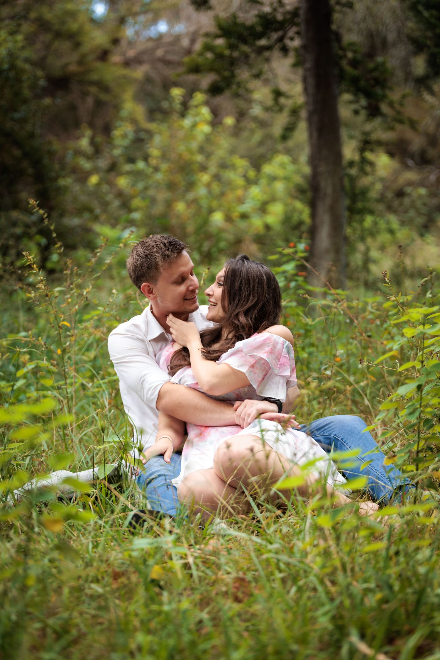 A romantic couple shares tender moments while lying in tall grass and wildflowers in a wooded outdoor setting.