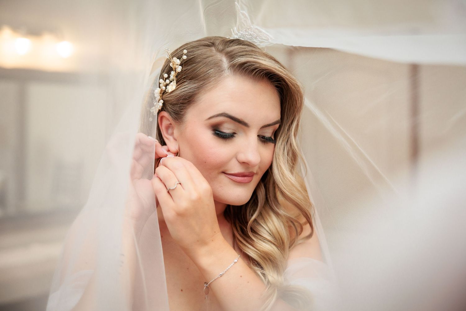 Bride in a veil adjusting her earring, wearing bridal hair accessories and elegant makeup on her wedding day.