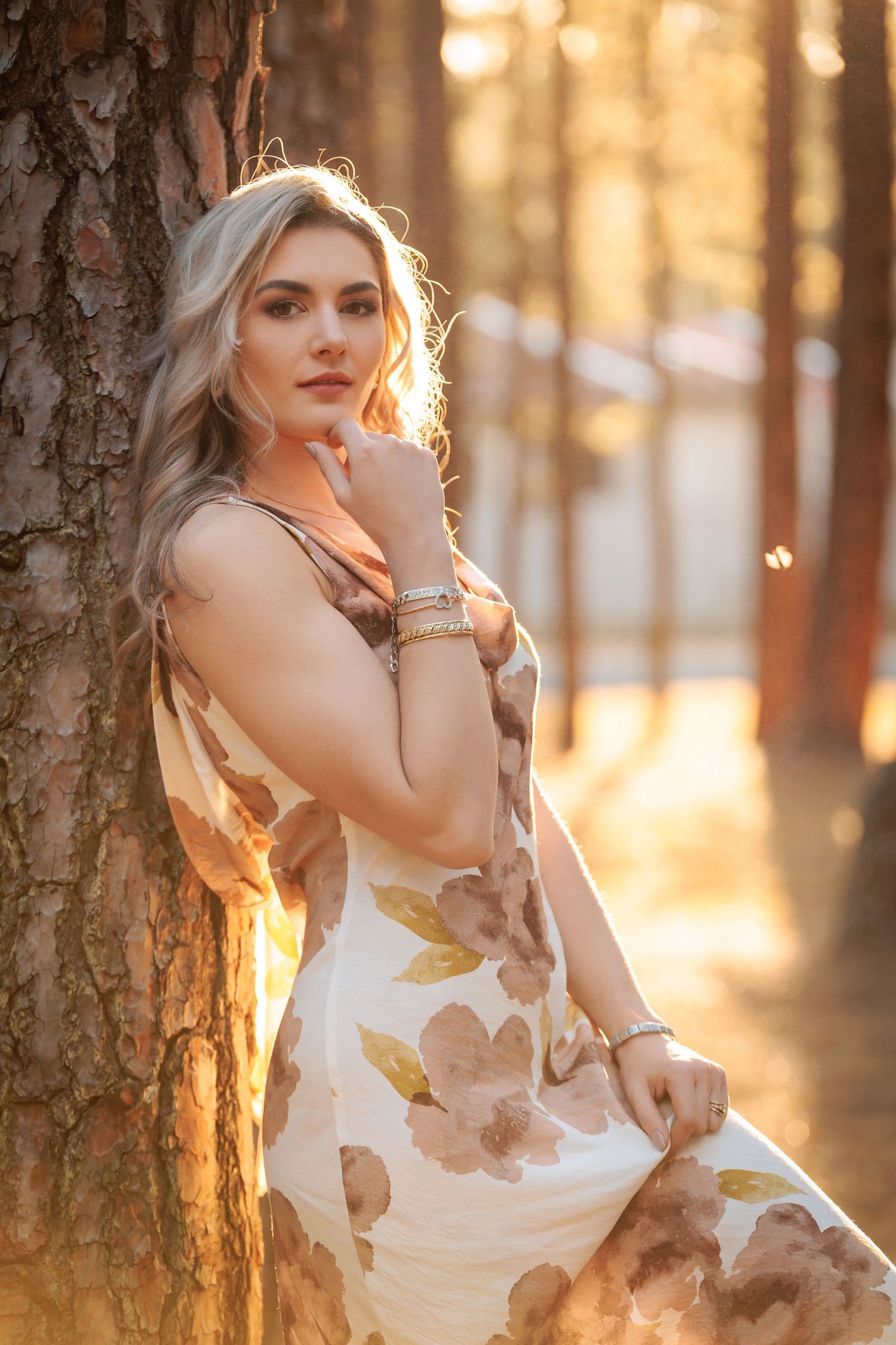 Woman in floral dress poses in sunlit forest during golden hour creating dreamy atmospheric portraits.