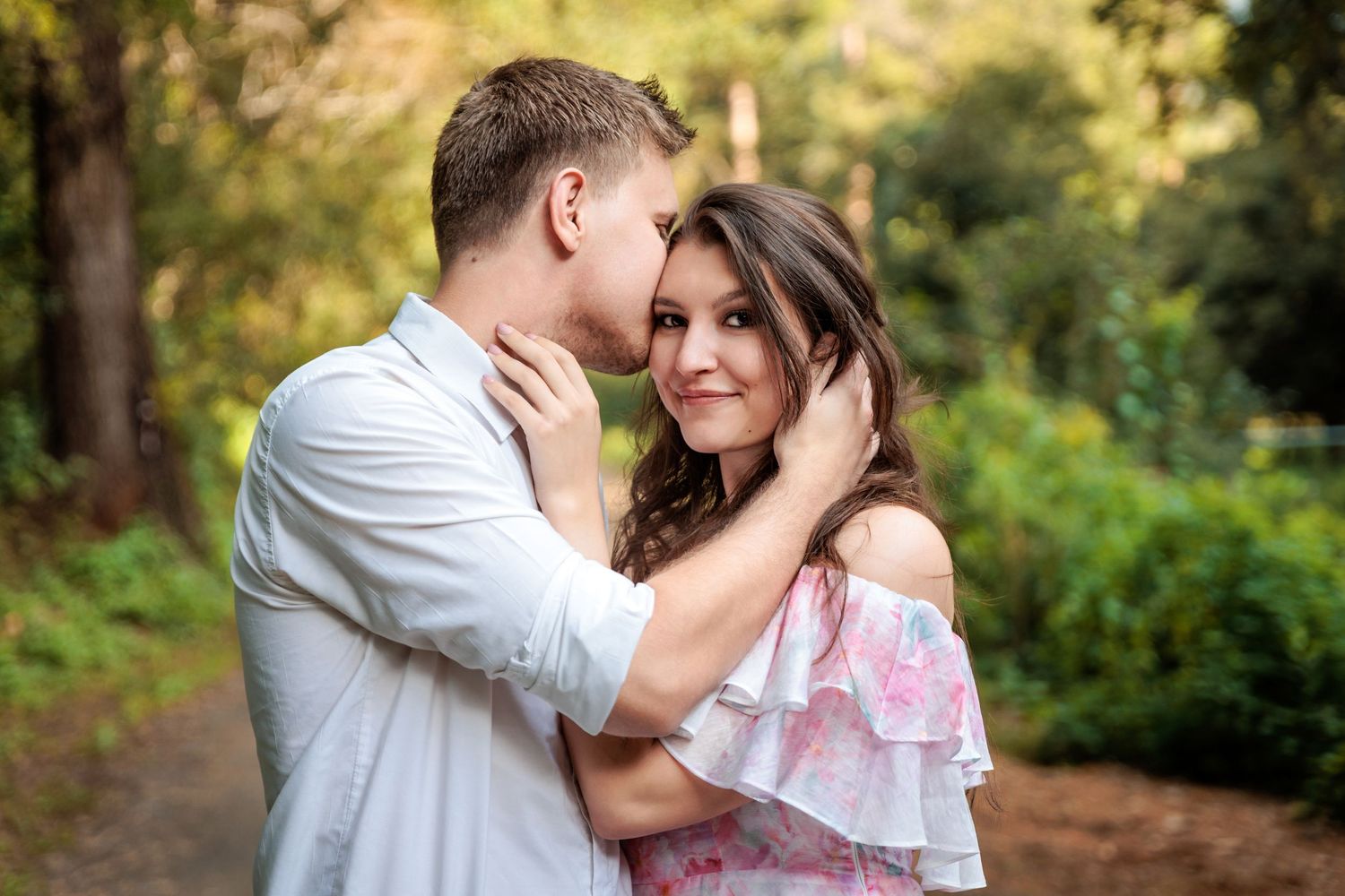 A romantic couple embraces outdoors during a photo session in a sunlit wooded setting.