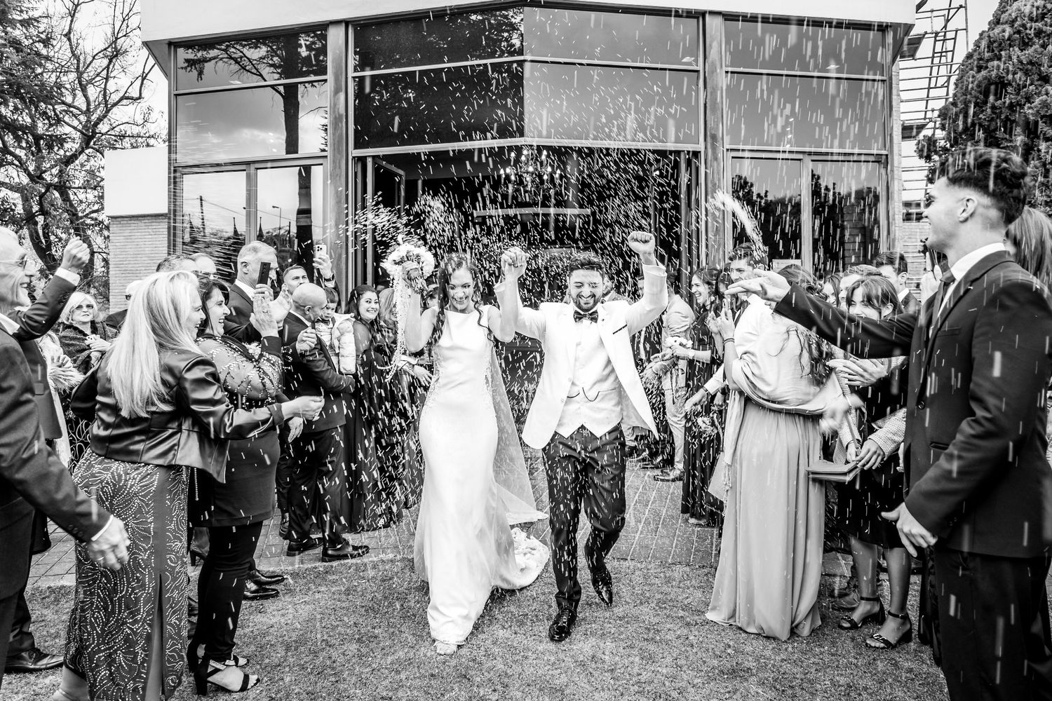 Black and white photo of a joyful bride and groom exiting their wedding ceremony as guests shower them with confetti.