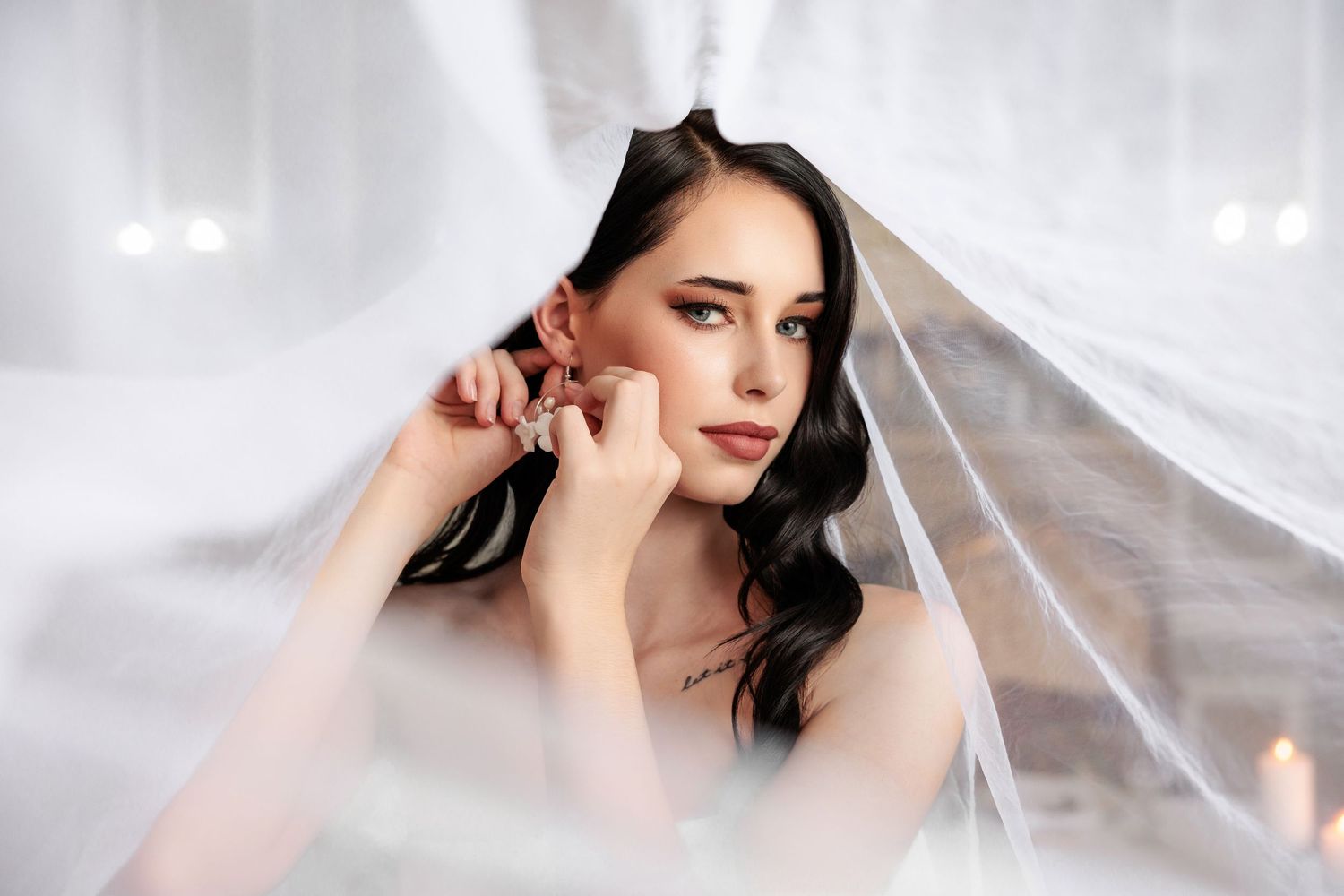 A beautiful dark-haired bride adjusts her earring under a flowing white veil in an elegant bridal portrait.