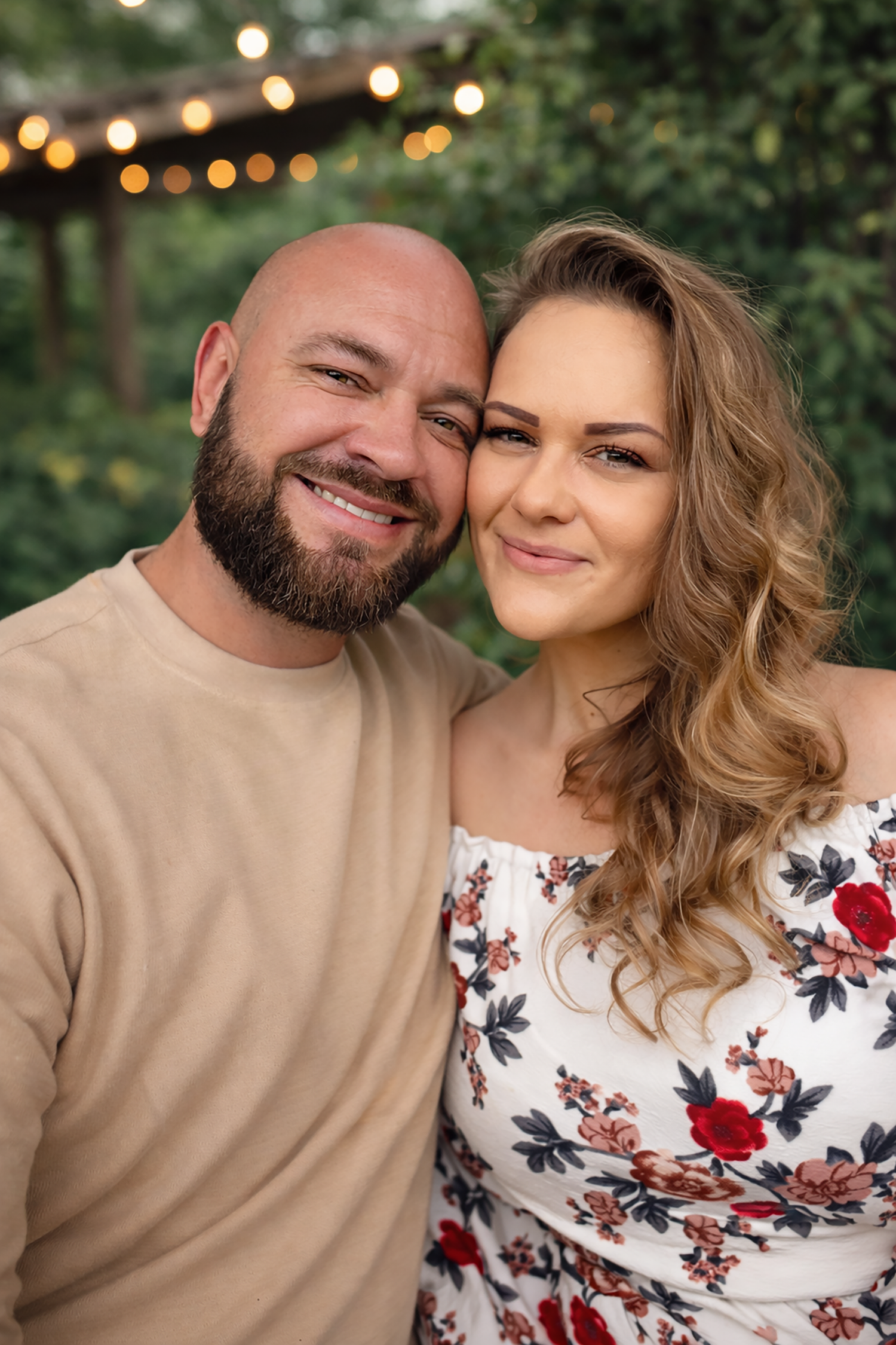 A couple poses outdoors with string lights in background wearing tan and floral outfits.