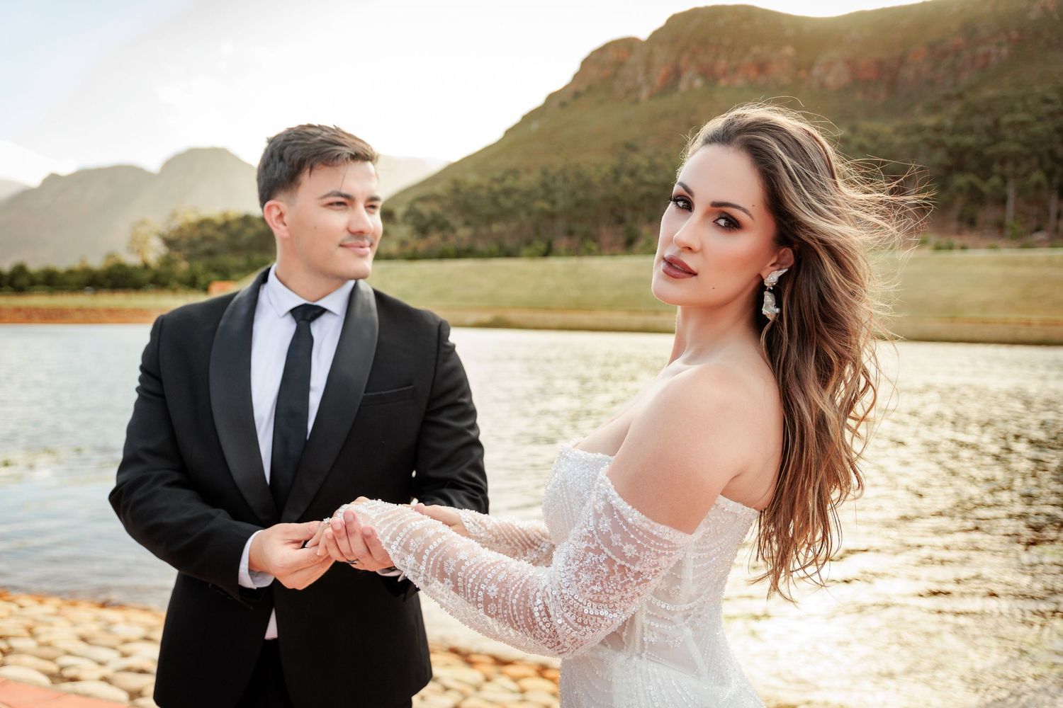 A couple in wedding attire walks along a scenic beach at sunset with mountains in the background.