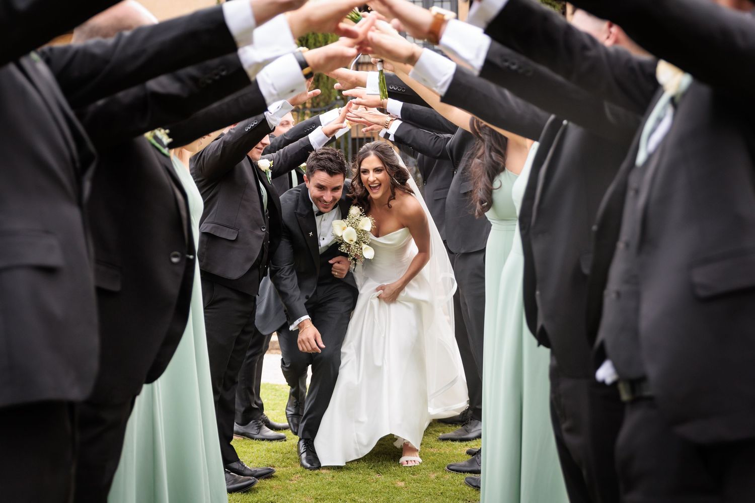 Wedding party members raise their arms to form a celebratory tunnel arch for the newlyweds to pass through outdoors.
