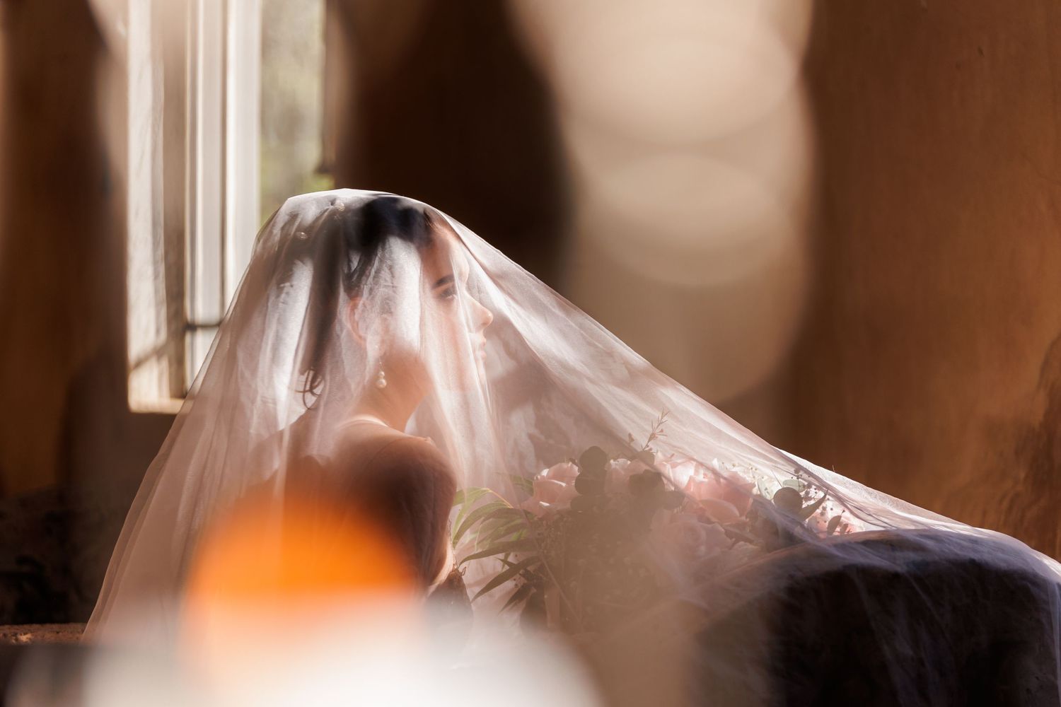 Bride covered by a flowing veil near a sunlit window, creating a dreamy, romantic wedding atmosphere.
