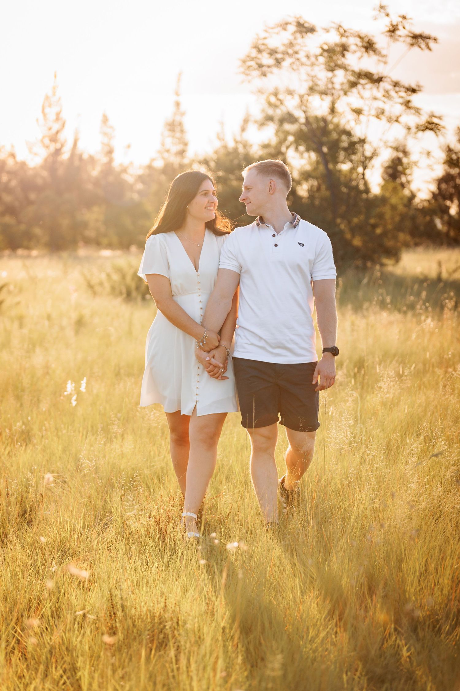 Couple walking hand in hand through a golden sunlit meadow at sunset wearing white and dark casual outfits.