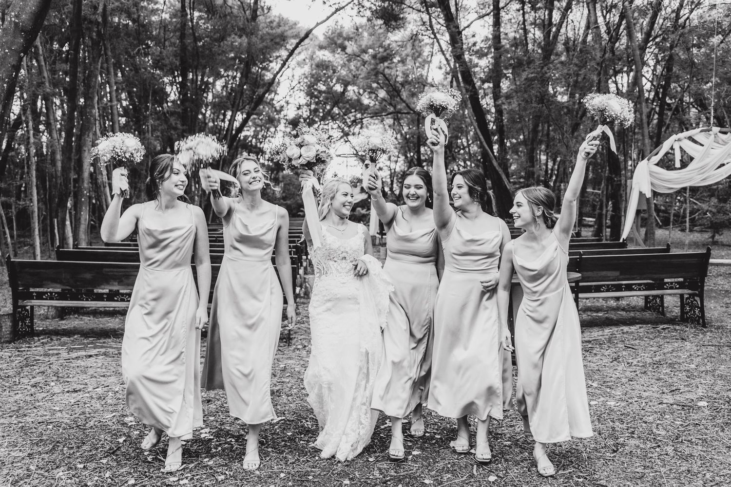 Wedding party in light colored dresses walking together through a wooded outdoor setting.