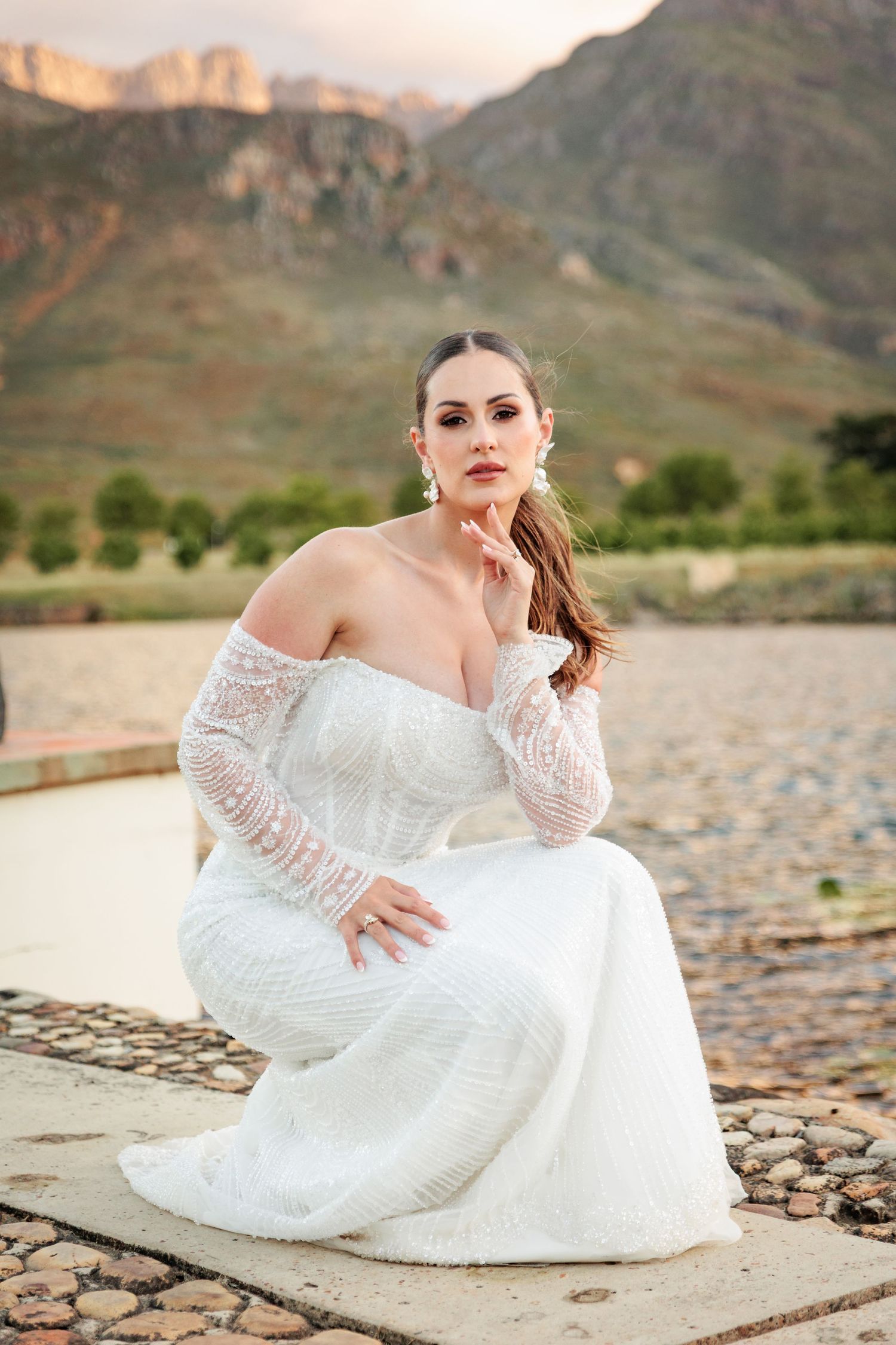 A person in a white lace wedding dress poses gracefully on rocky terrain with mountains in the background.
