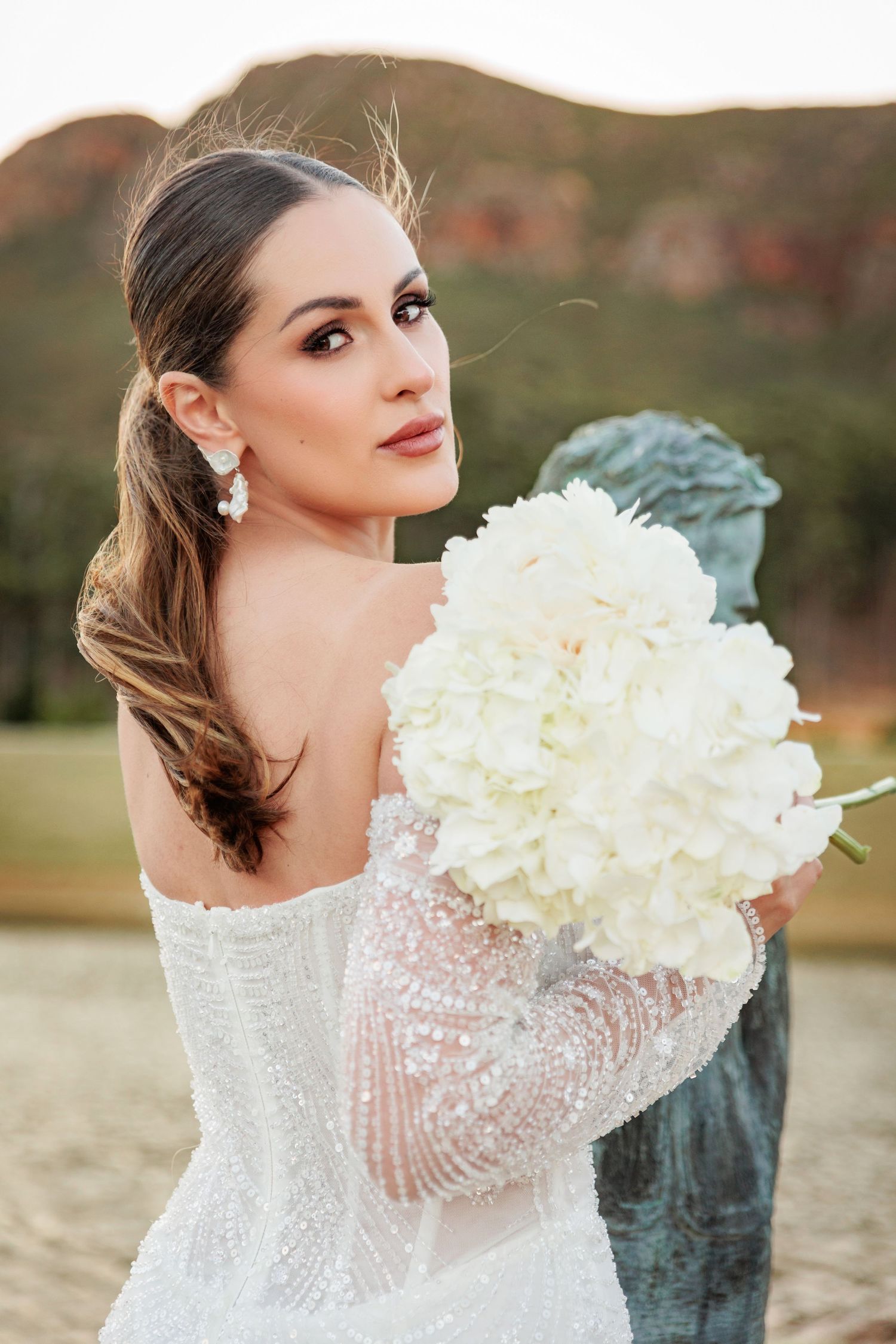 A bride in a lace sleeve wedding dress holds a bouquet of white hydrangeas in a romantic outdoor setting.
