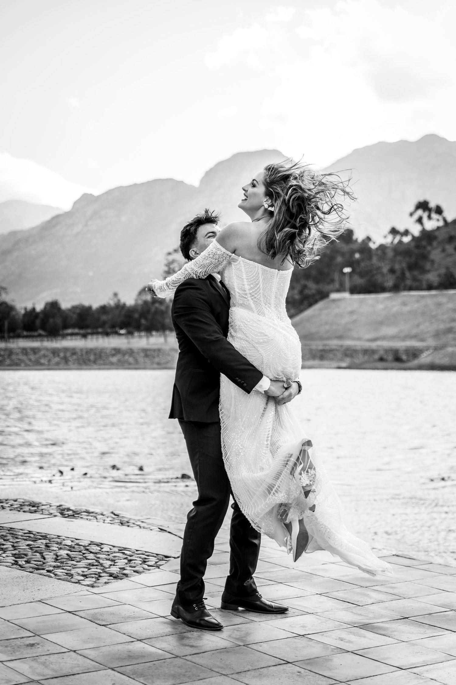 A romantic wedding couple embraces as wind blows the bride's veil on a stone patio near mountains and water.