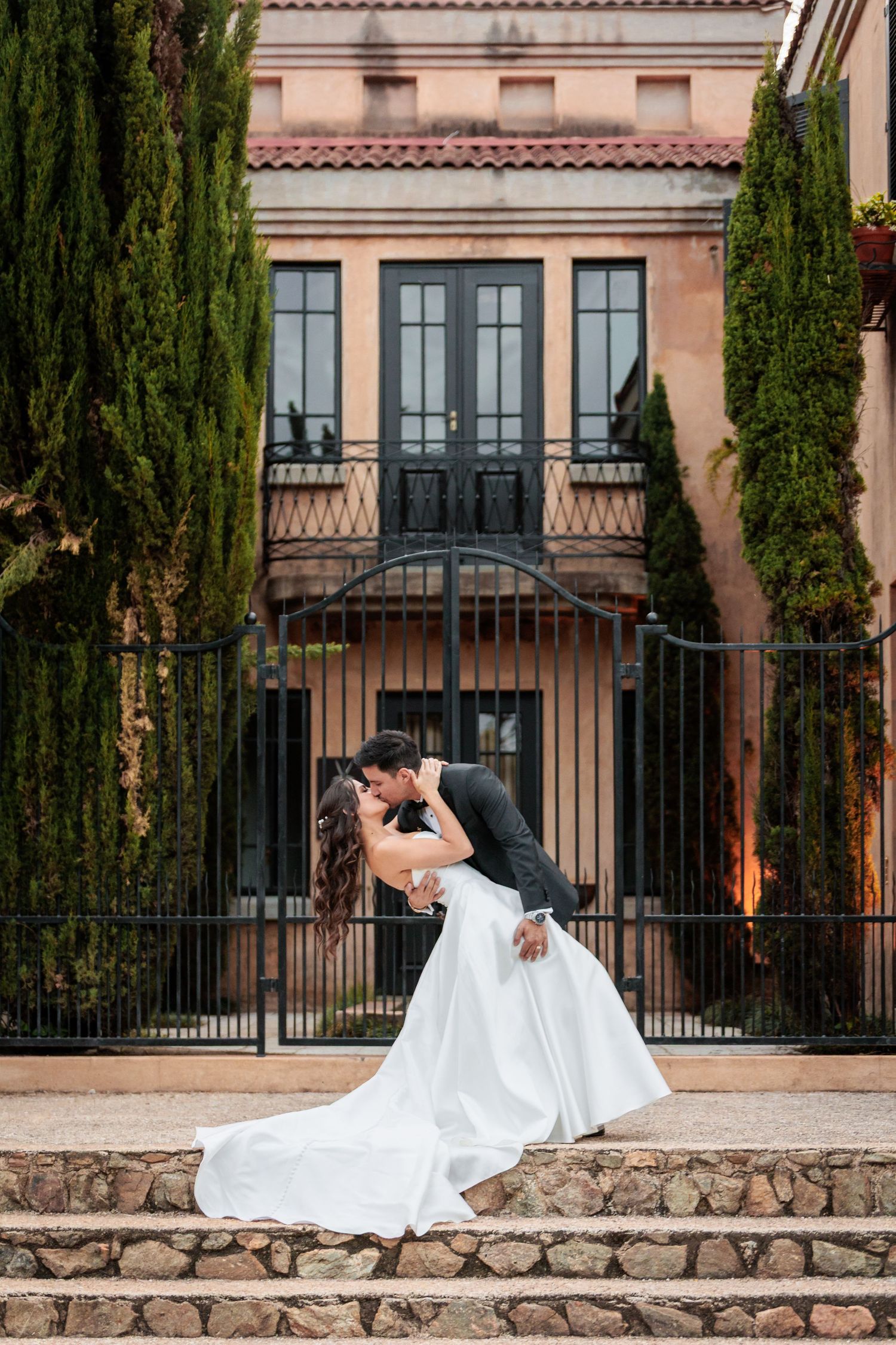 A romantic couple in wedding attire shares intimate poses on a stone wall against an elegant Mediterranean-style backdrop.