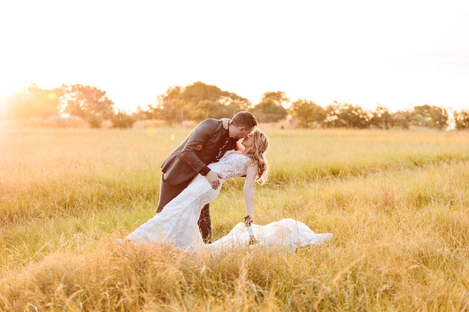 A romantic couple in wedding attire share an intimate moment in a golden sunlit field at sunset.