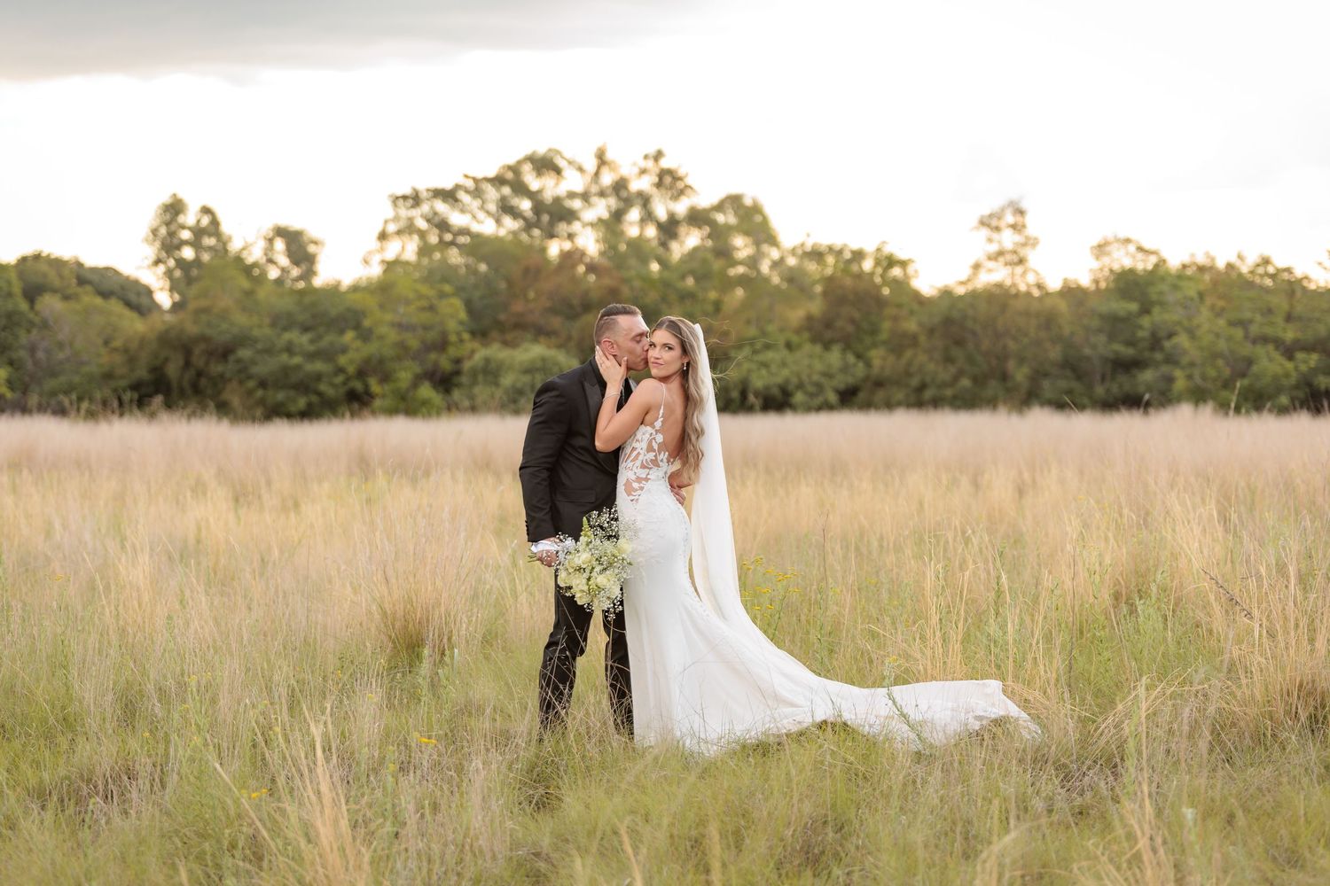 A bride and groom share a romantic kiss in a golden grass field at sunset, surrounded by lush green trees.