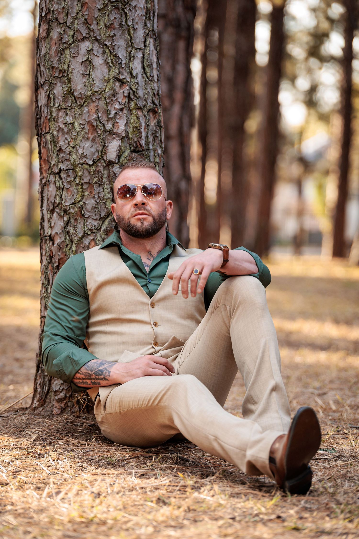A person in a green shirt and khaki pants reclines against a tree trunk in a forest setting during autumn.