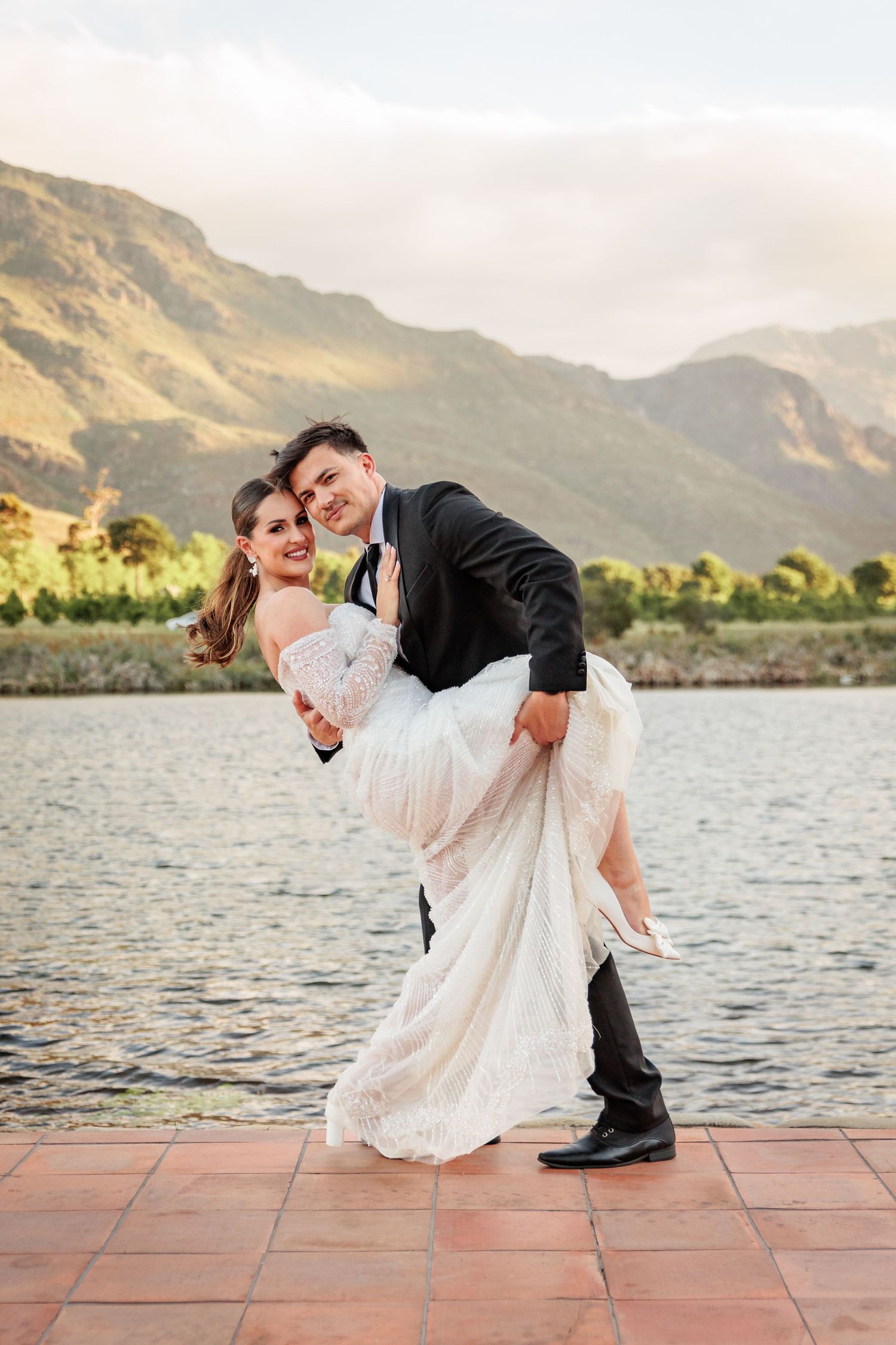 A romantic dip pose sequence between a couple in formal wedding attire by a lakeside mountain backdrop at sunset.