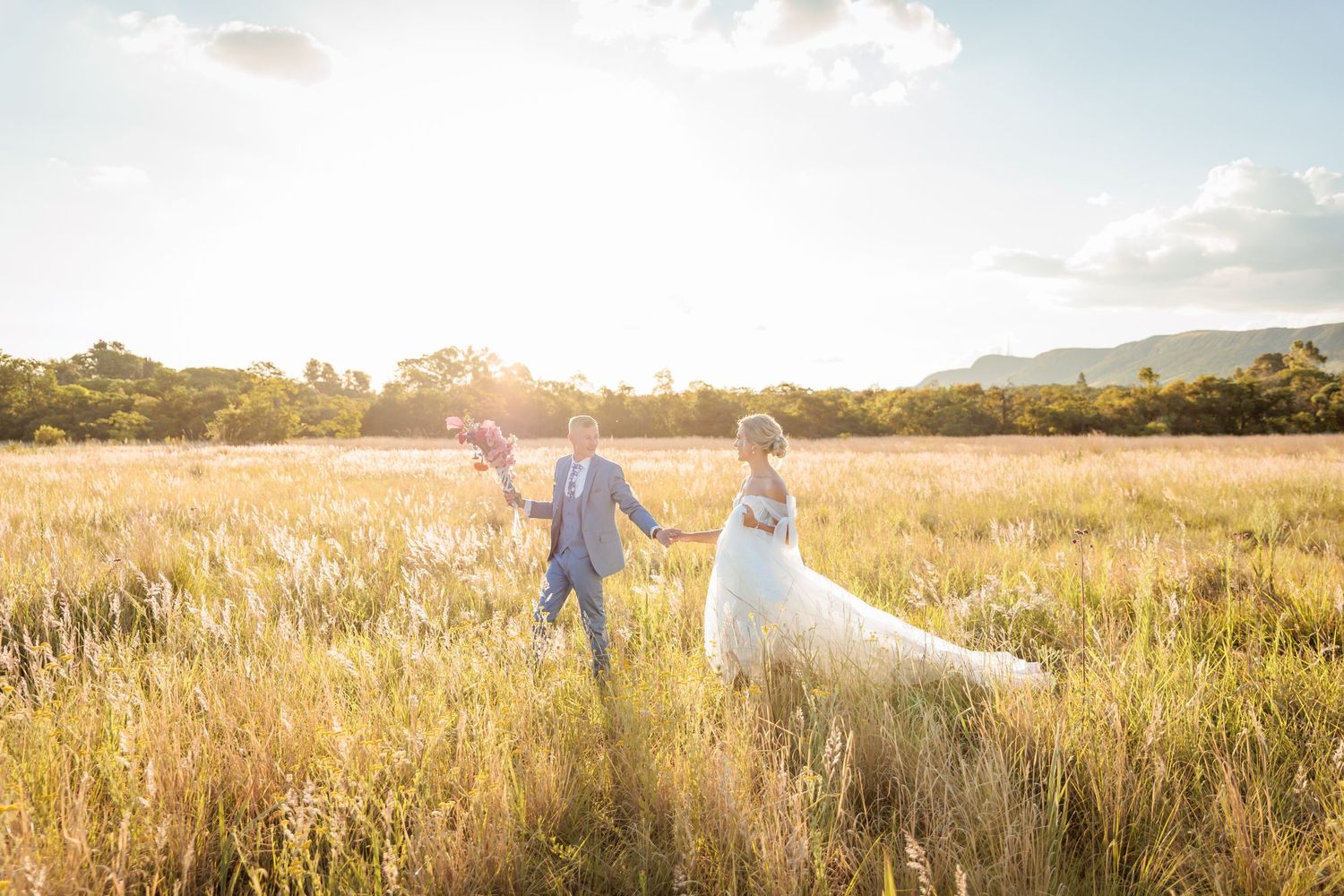 Bride and groom walking hand in hand through golden sunlit meadow with mountains in background on wedding day.