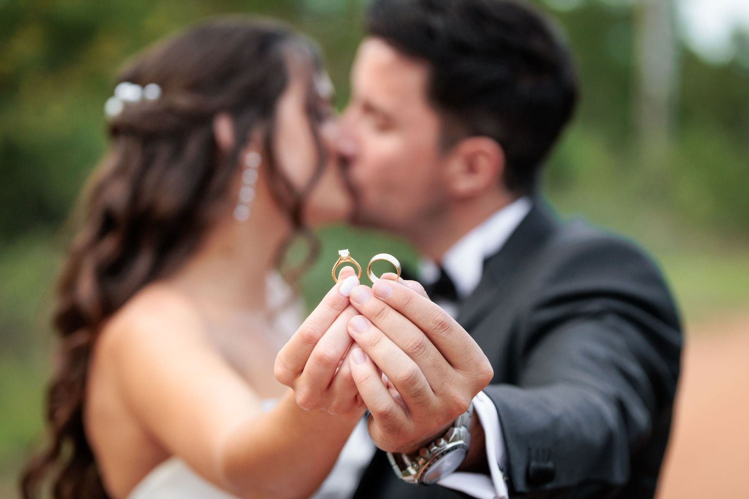 Wedding couple holds up gold rings while sharing a romantic kiss in an outdoor garden setting.