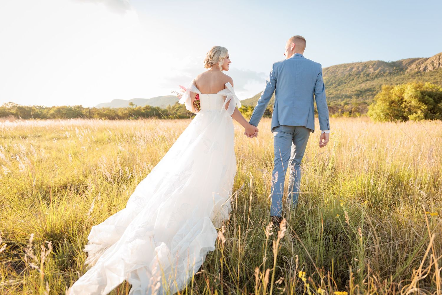 Bride and groom walking hand in hand through golden meadow with mountain backdrop on sunny wedding day.