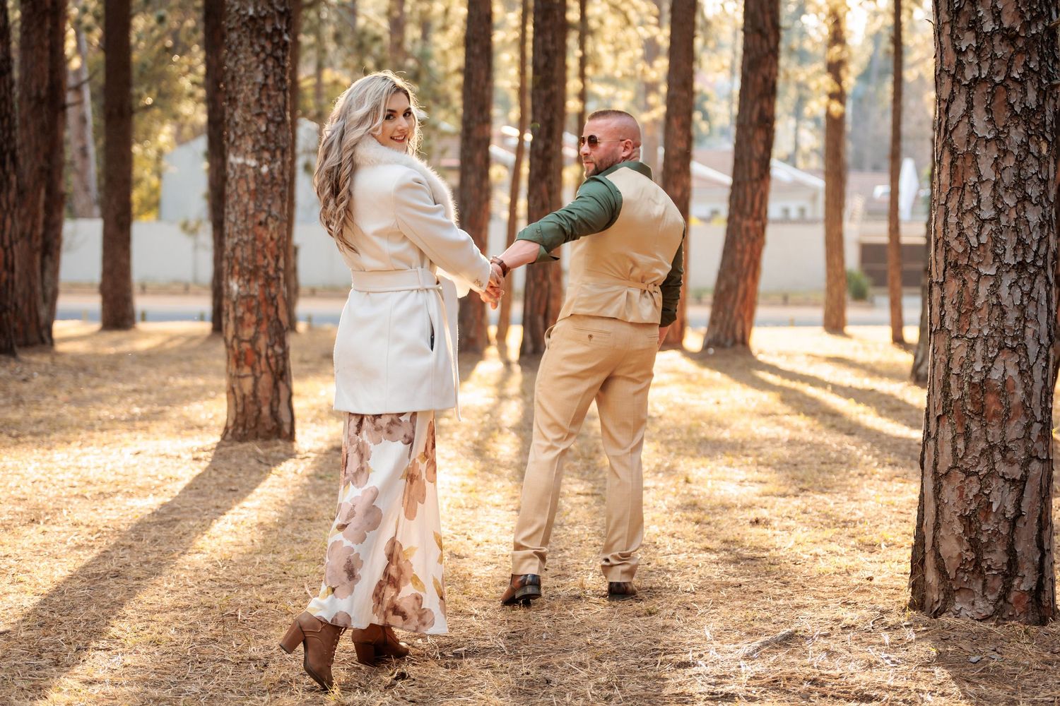 A couple in white and tan outfits share a romantic moment while walking through a sunlit forest.
