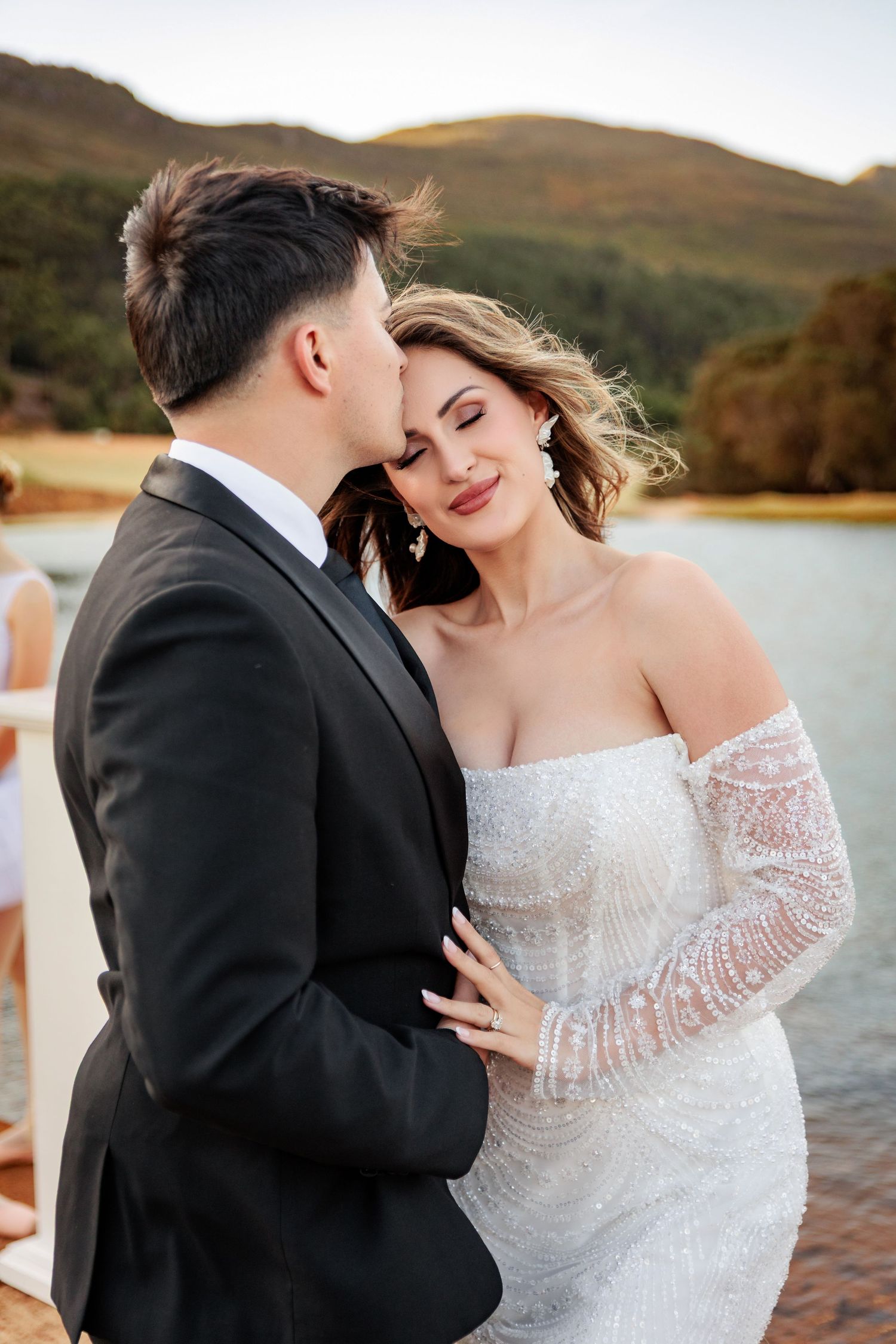 A couple in wedding attire embraces by a scenic lake during sunset, with mountains in the background.