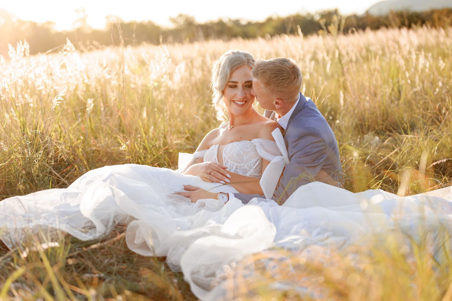 A couple in wedding attire sits together in a sunlit field of tall golden grass during sunset.