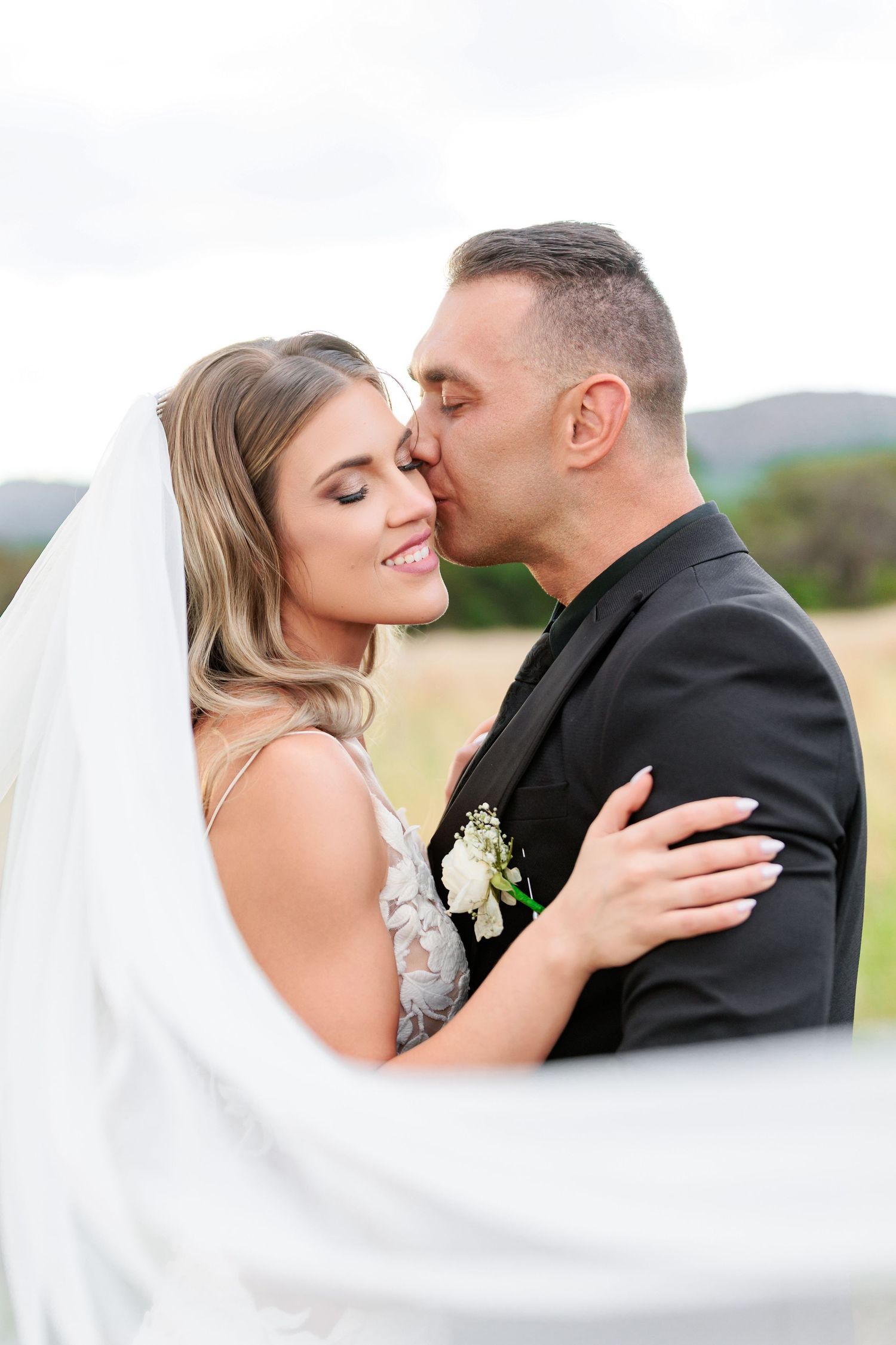 A romantic wedding couple shares tender kisses in an outdoor setting with flowing white veil in the foreground.
