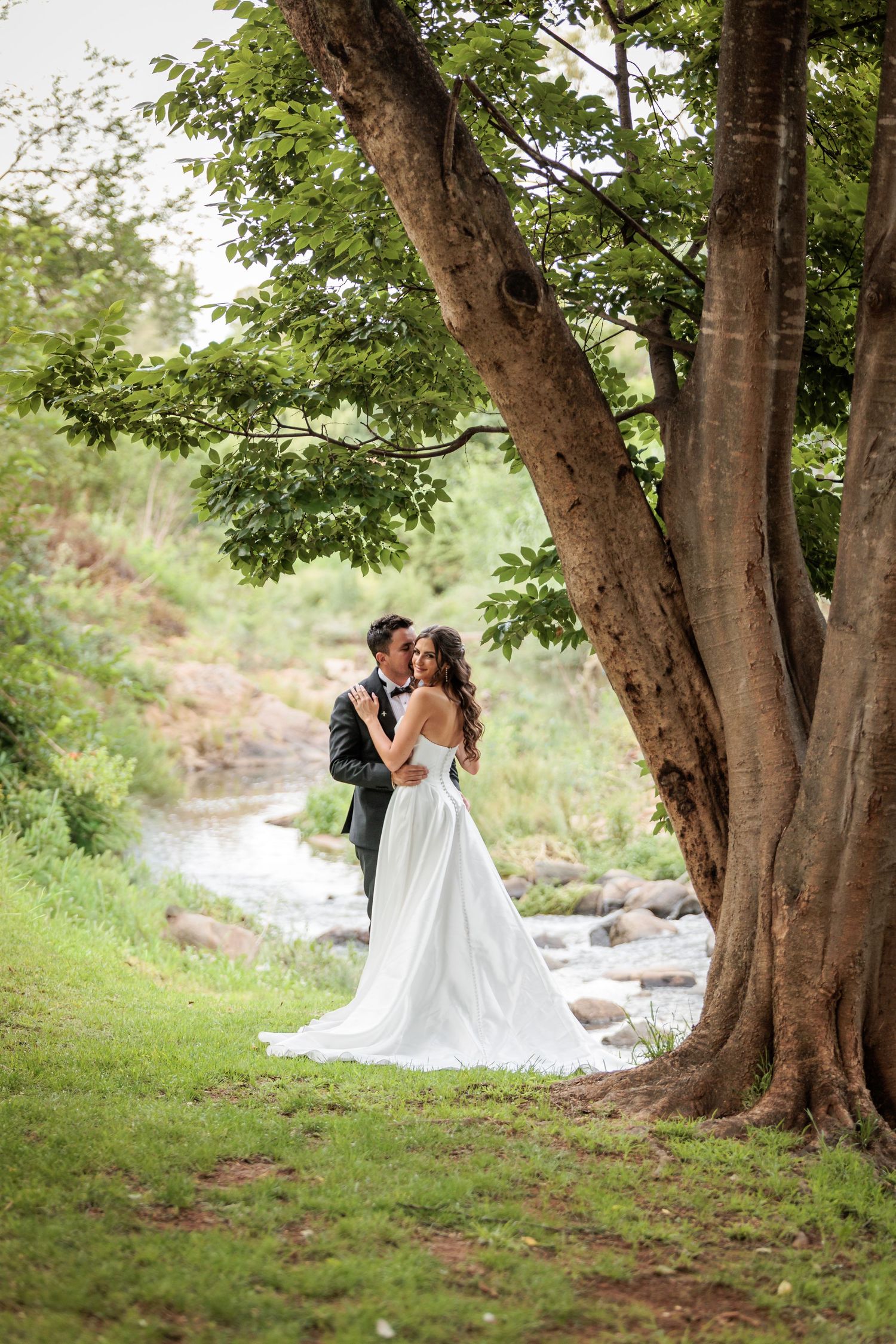 A romantic wedding couple embraces under a canopy of tall trees beside a flowing stream in a natural outdoor setting.