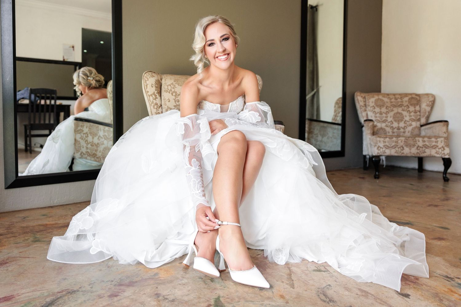 A bride in a white wedding gown and veil sits gracefully on a stone floor in an elegant indoor setting.