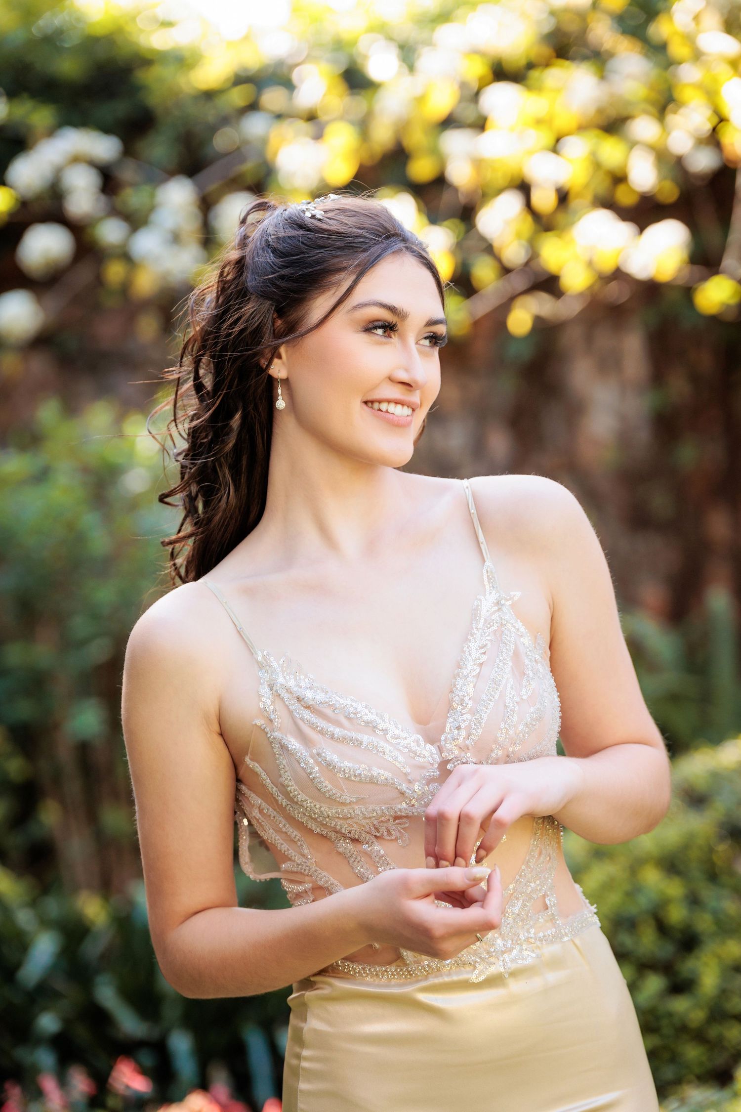 A person in a beige lace dress poses outdoors with sunlit foliage in the background during golden hour.