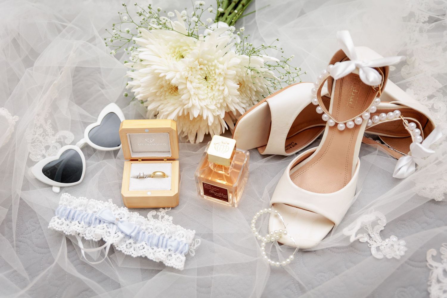 Wedding accessories including white flowers, shoes, and jewelry laid out on light fabric.