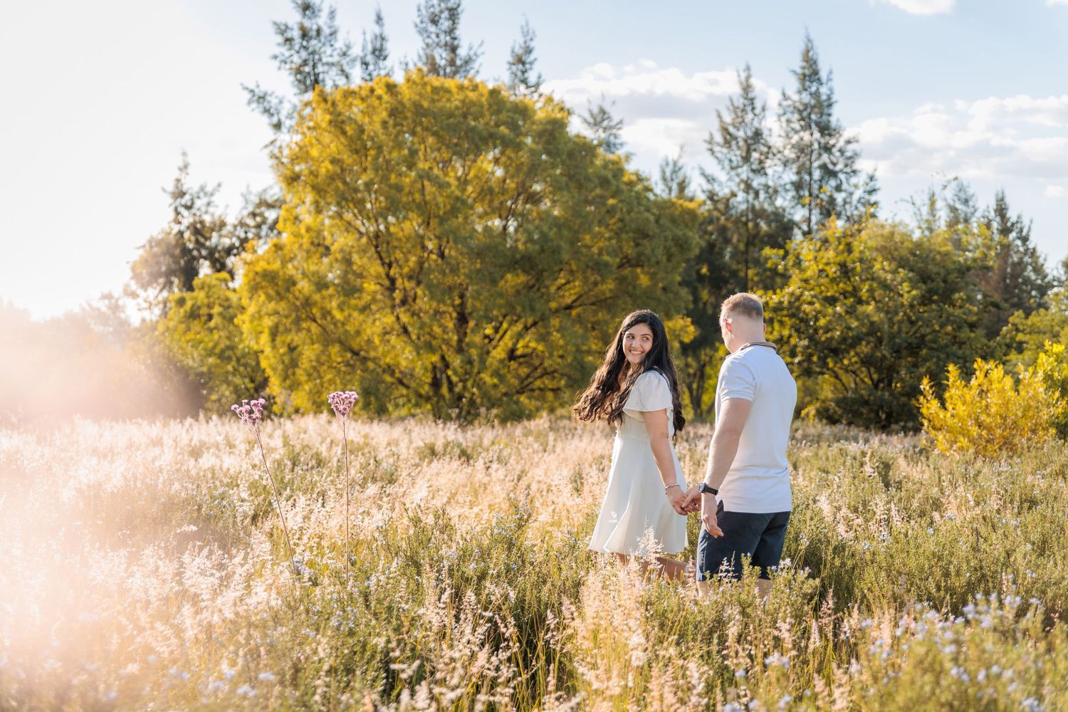 A couple stands together in a sunlit meadow surrounded by golden autumn trees and tall grass at sunset.